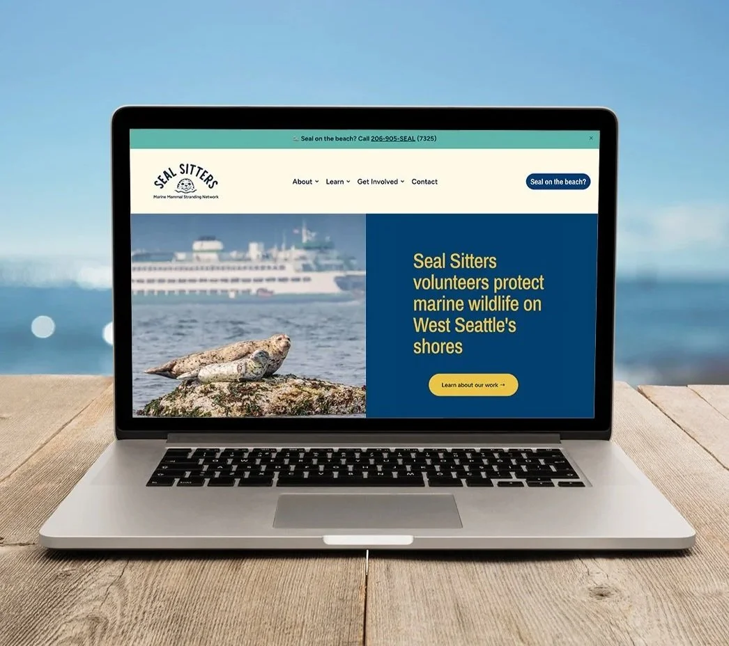 A laptop on a wooden table displaying a website for Seal Sitters, an organization that protects marine wildlife on West Seattle's shores, with a background of ocean and a ship.