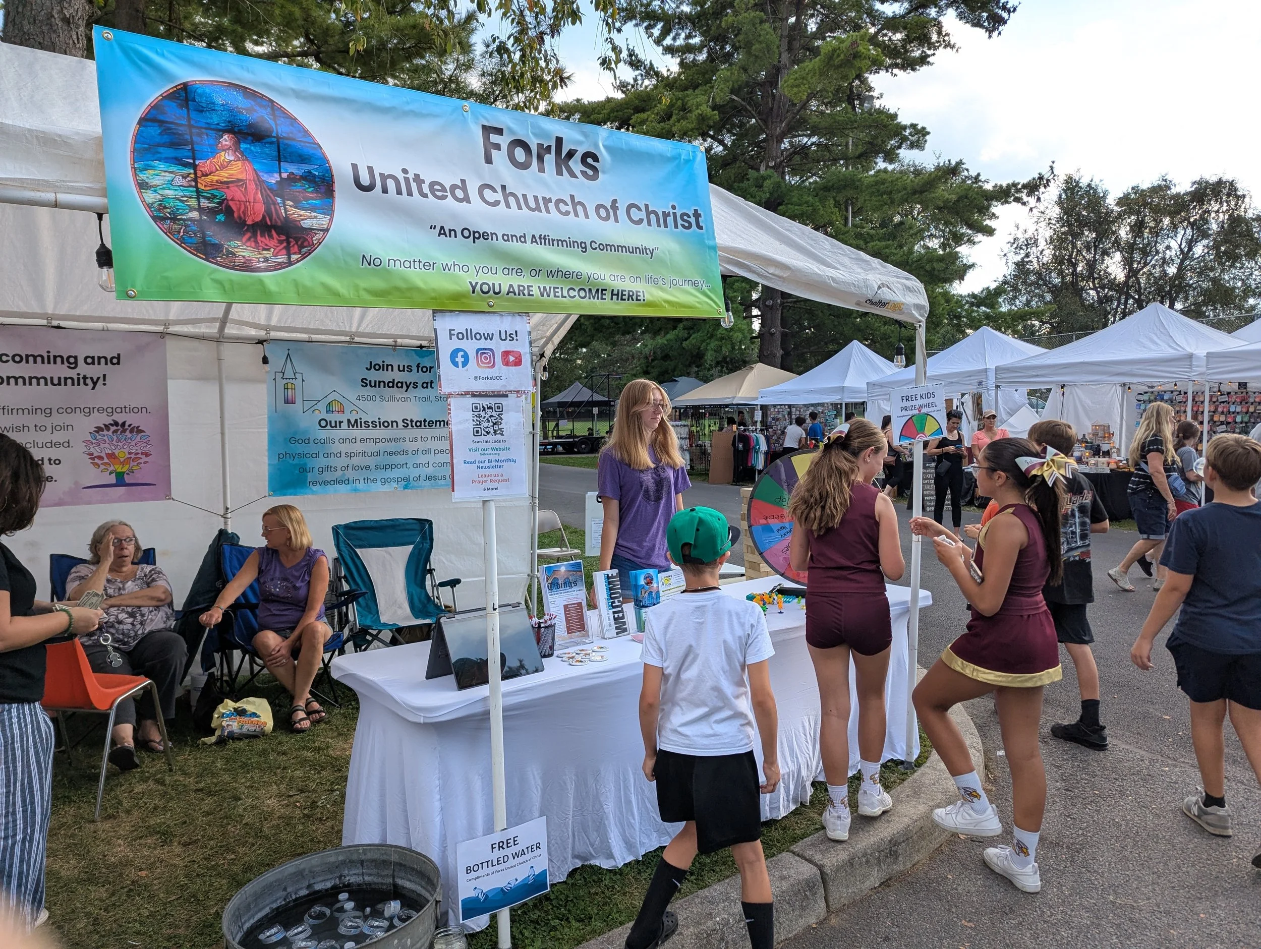 A community outdoor fair with booths, including one for Forks United Church of Christ, featuring a spinning wheel game and informational materials, with people of various ages engaging at the booths and walking around.