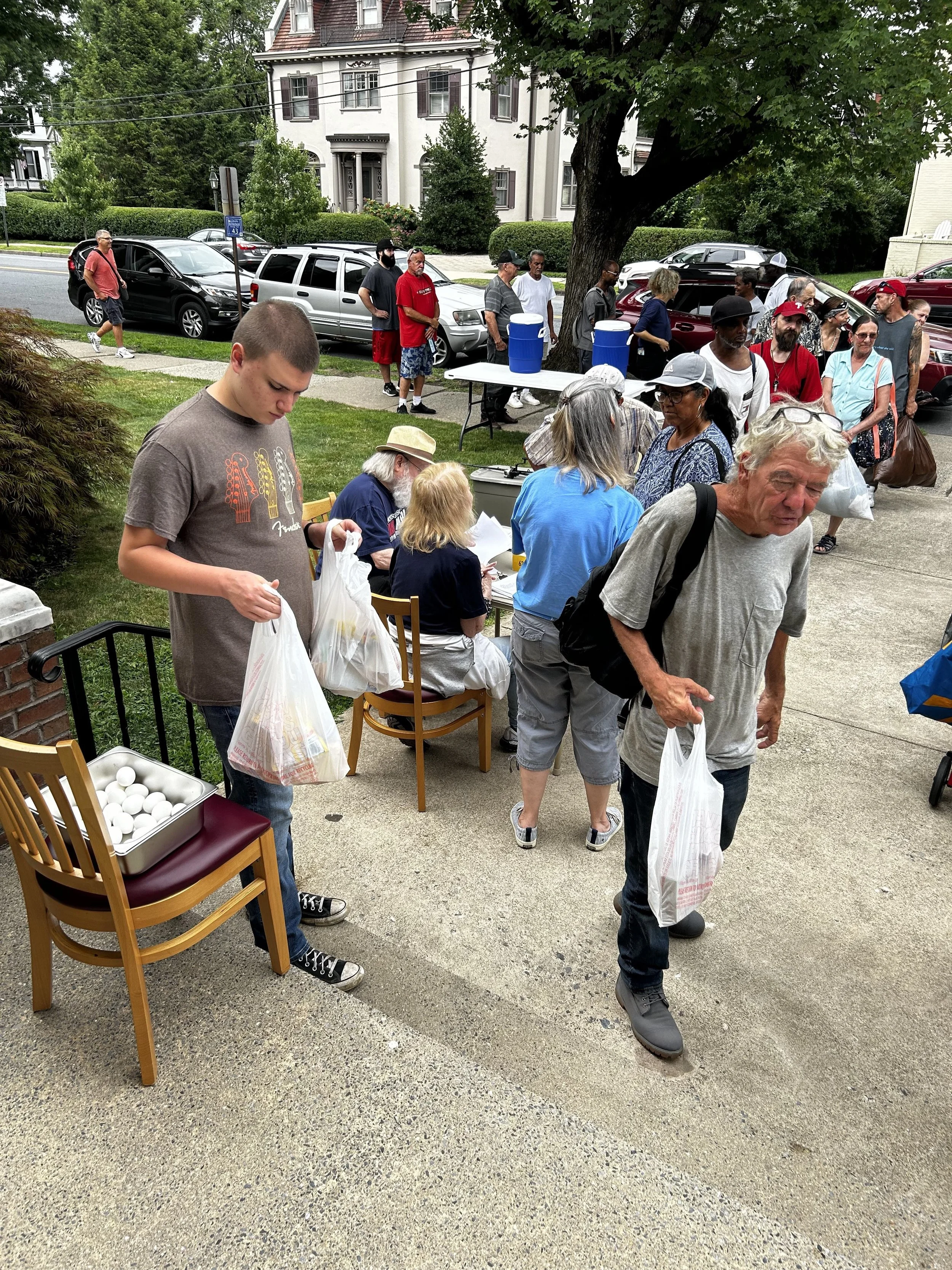 People standing in a line outside a house, some holding bags, with tables set up for distribution, including a tray of eggs on a chair, on a sidewalk in a residential neighborhood.