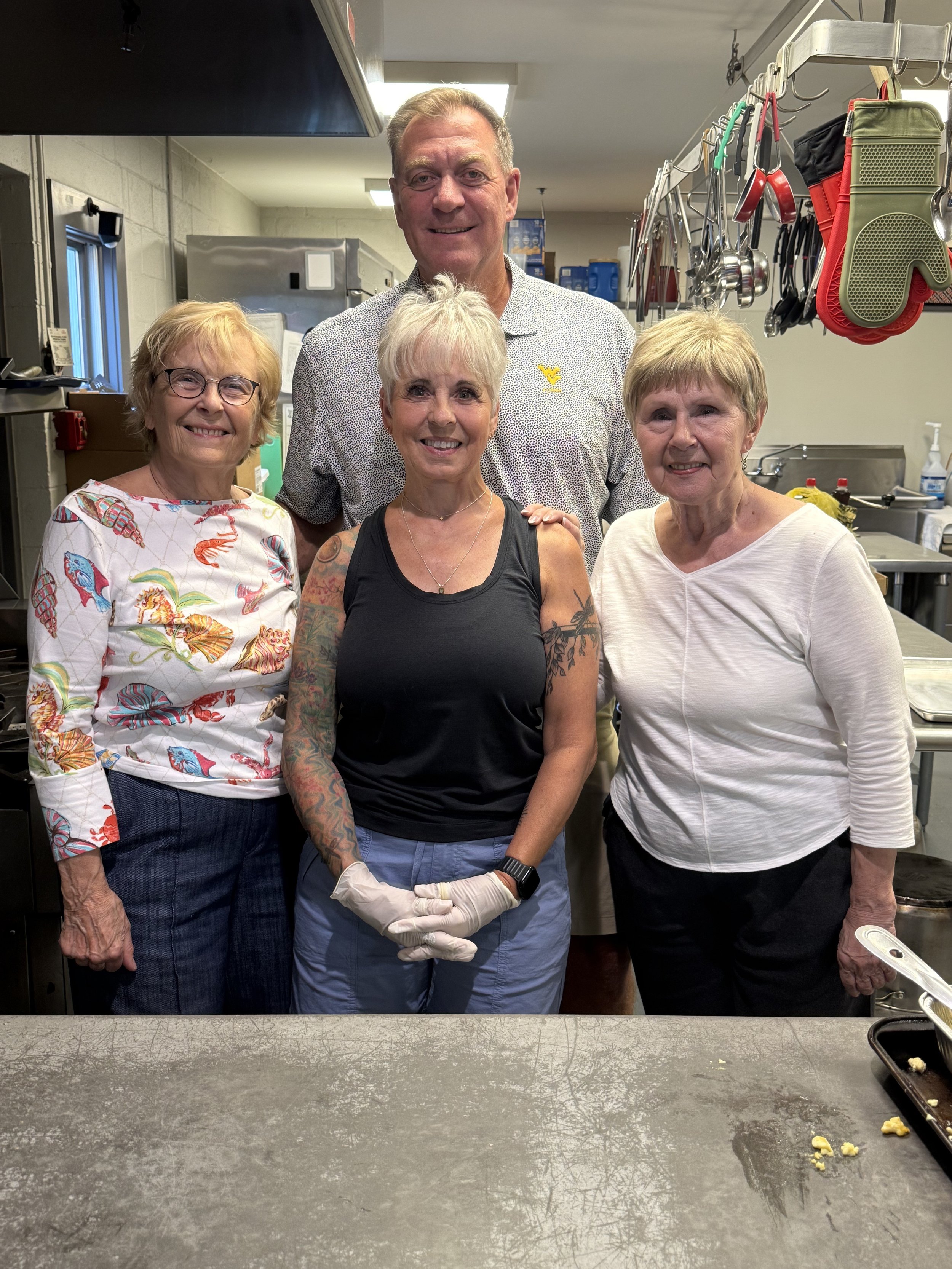 Four adults posing together in a kitchen, one woman with tattoos and gloves, others smiling, behind a cooking area with utensils. The group appears friendly and cheerful.