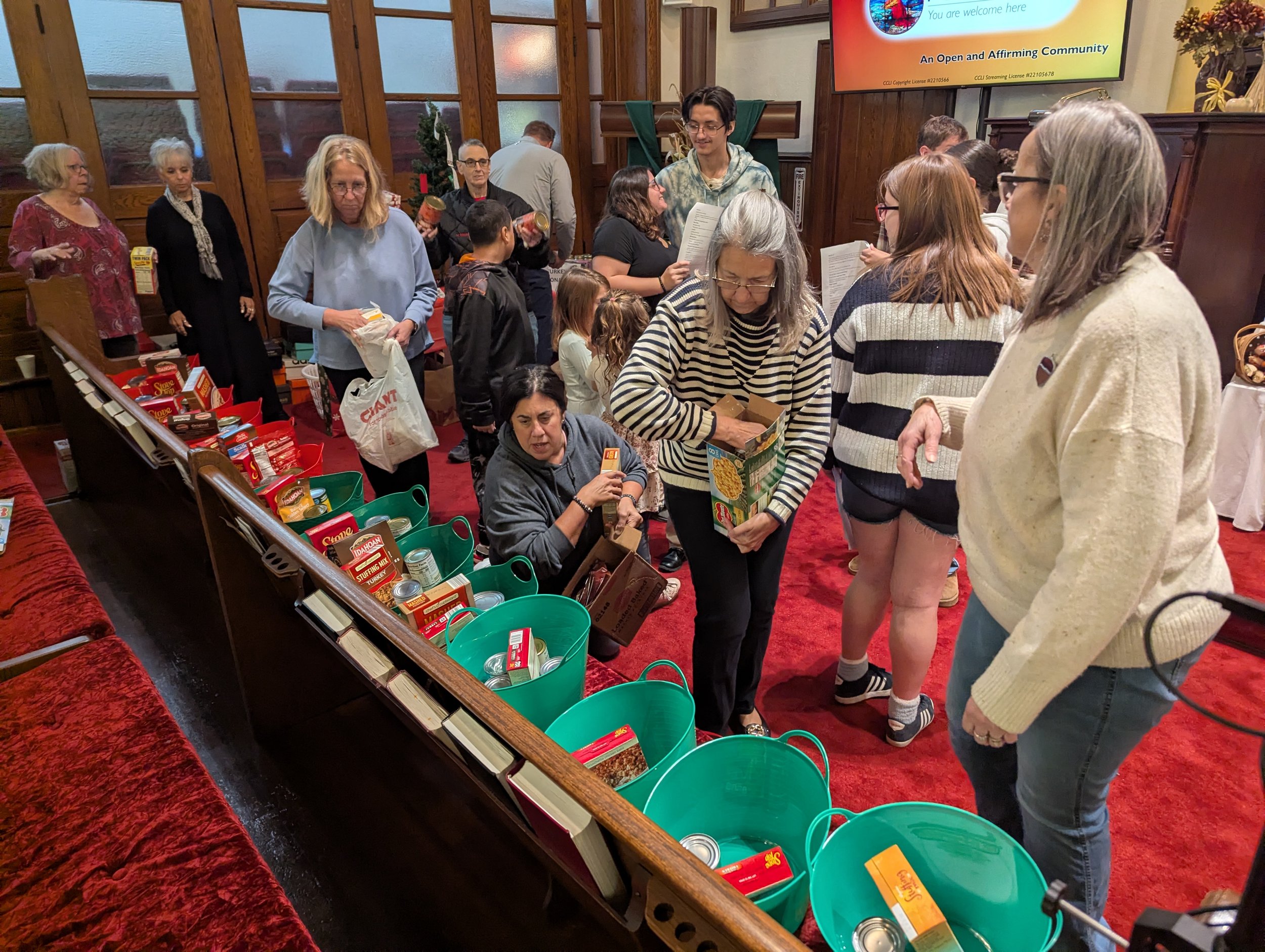 People participating in a food drive or donation event, showing a variety of individuals in a room with wooden paneling and red carpet, organizing and collecting canned goods and non-perishable food items.
