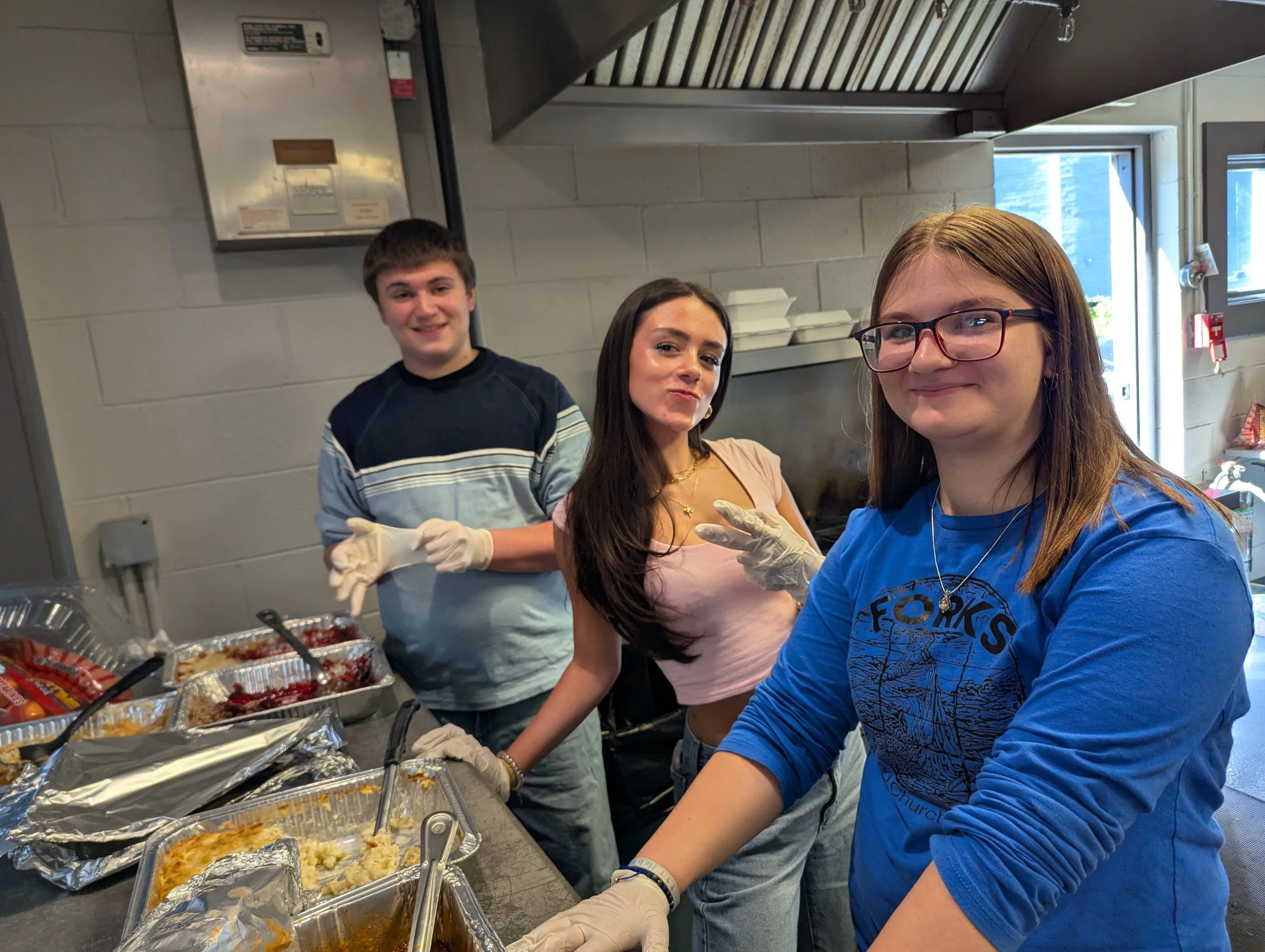 Three teenagers in a kitchen serving food, wearing gloves, smiling, and making peace signs. The girl in front has glasses and a blue shirt, the girl in the middle has long dark hair and a pink shirt, the boy in the back has short hair and a striped s