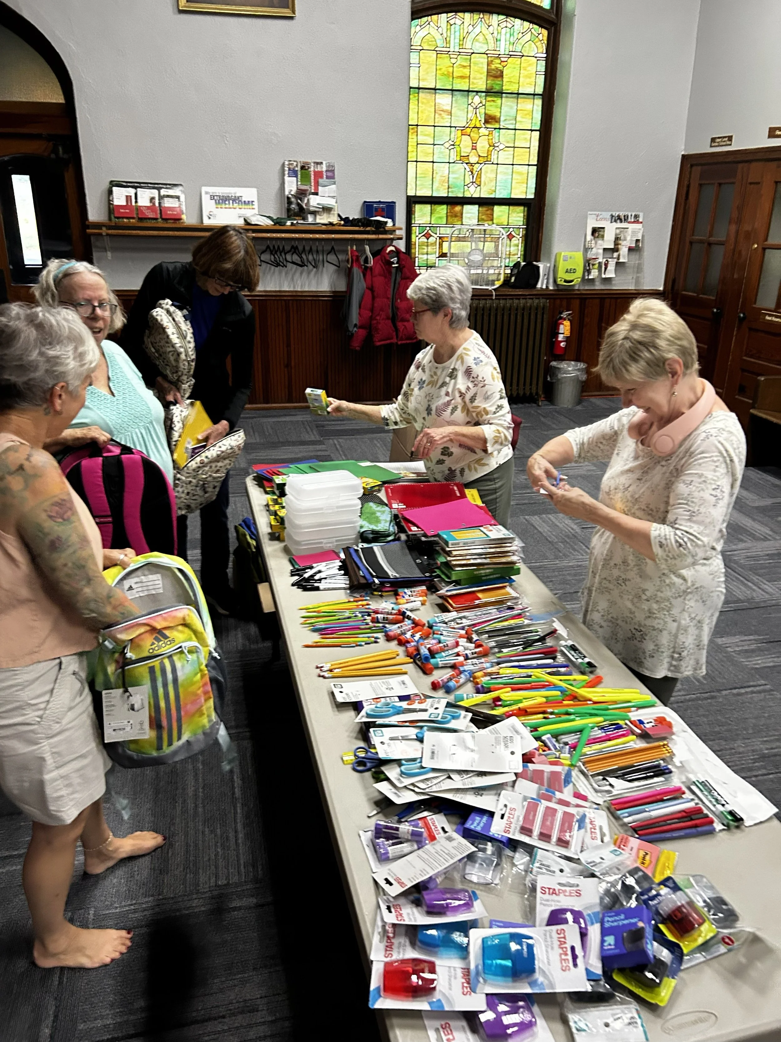 A group of people shopping for school supplies at a table with various pens, markers, scissors, and other office items in a room with stained glass windows.