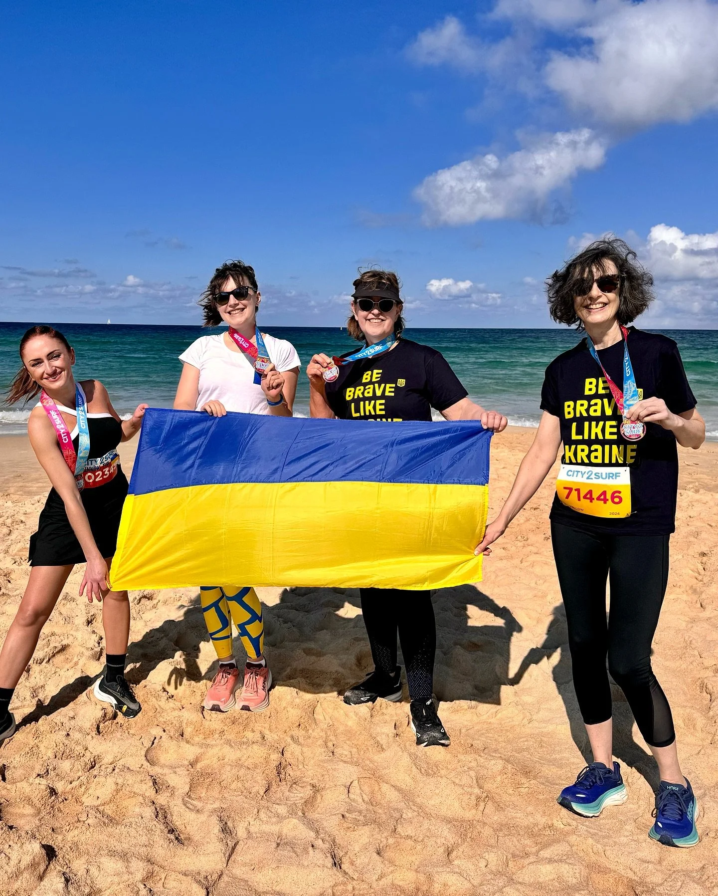 Four women standing on a sandy beach holding a Ukrainian flag and wearing medals, with the ocean and blue sky in the background.