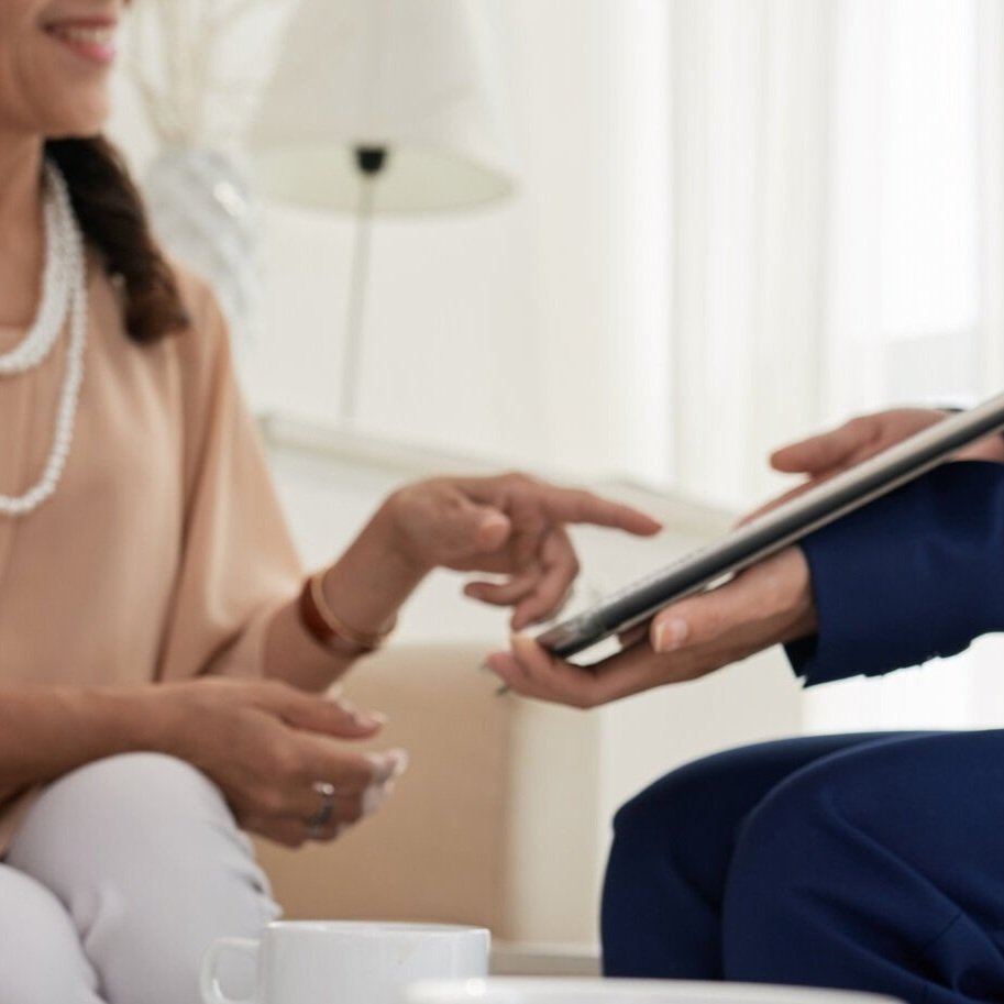 A woman wearing a necklace consults with a professional, who is handing her a tablet. The professional is dressed in a suit. The setting appears to be a consultation or appointment.