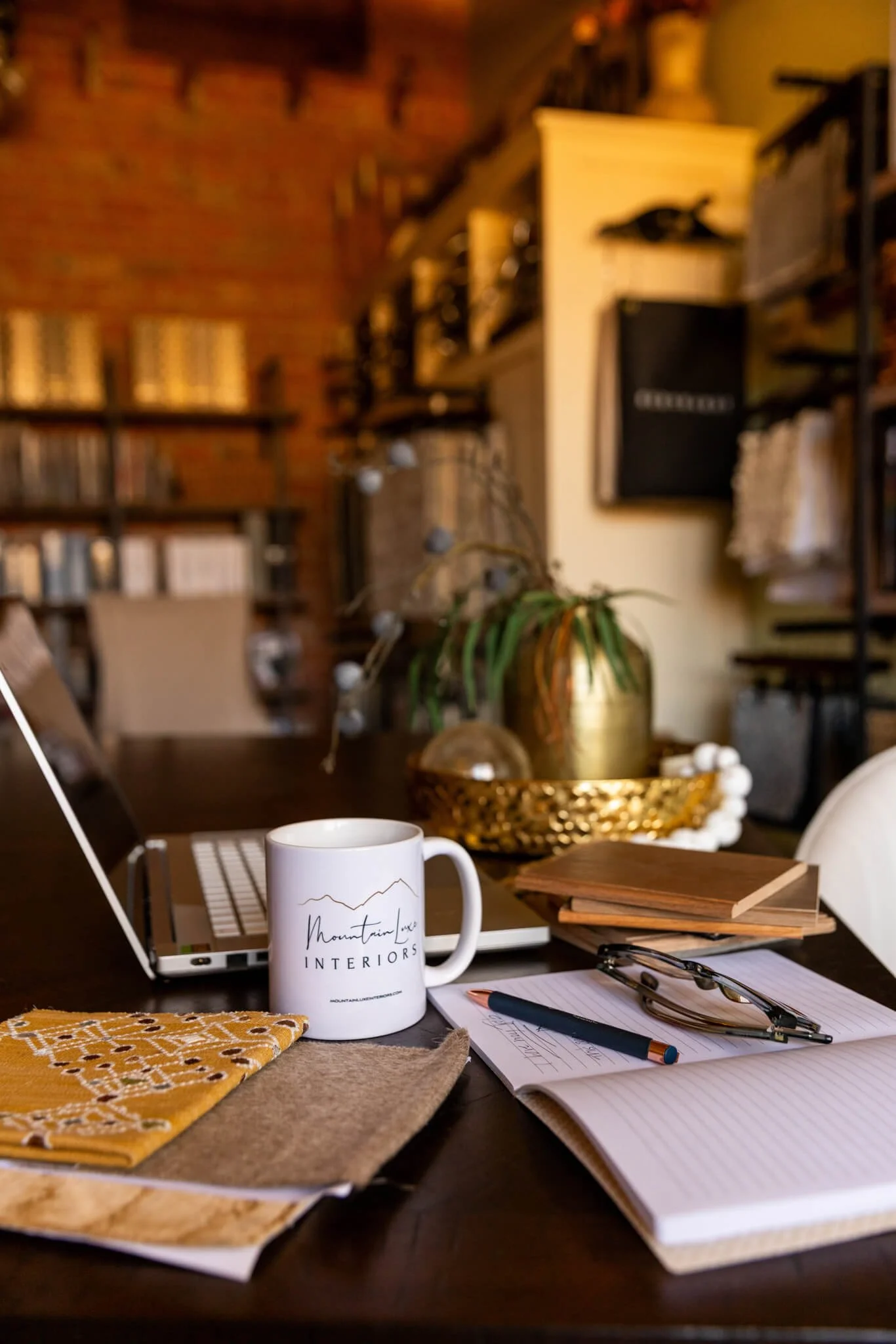 A desk with a white mug that has the logo "Mountain Luxe Interiors," a yellow and white patterned cloth, several notebooks, a pen, reading glasses, and a laptop. In the background, there are bookshelves, a potted plant, and wall decorations in a cozy interior space.