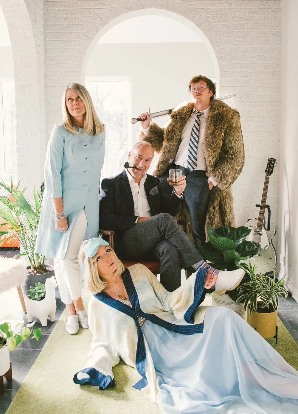 A group of five people posing in a bright, modern room with plants and guitars. One person is sitting and holding a drink, another standing with a fur coat and a sword, a woman standing in a light blue coat, a woman lying on the floor in a white and blue dress with a sleep mask, and a man with a headset and white shirt sitting on a chair.