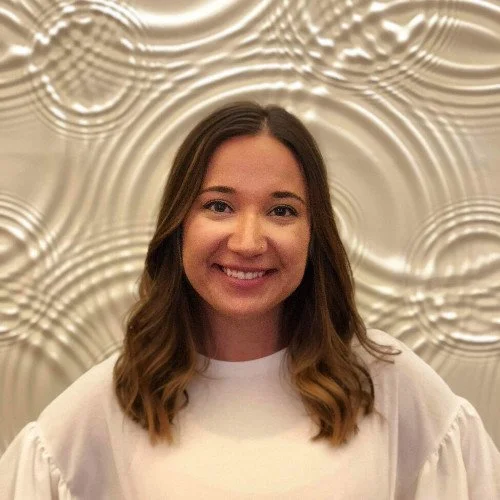 A young woman with shoulder-length brown hair, smiling, wearing a white top, standing in front of a textured, metallic, silver-colored wall.