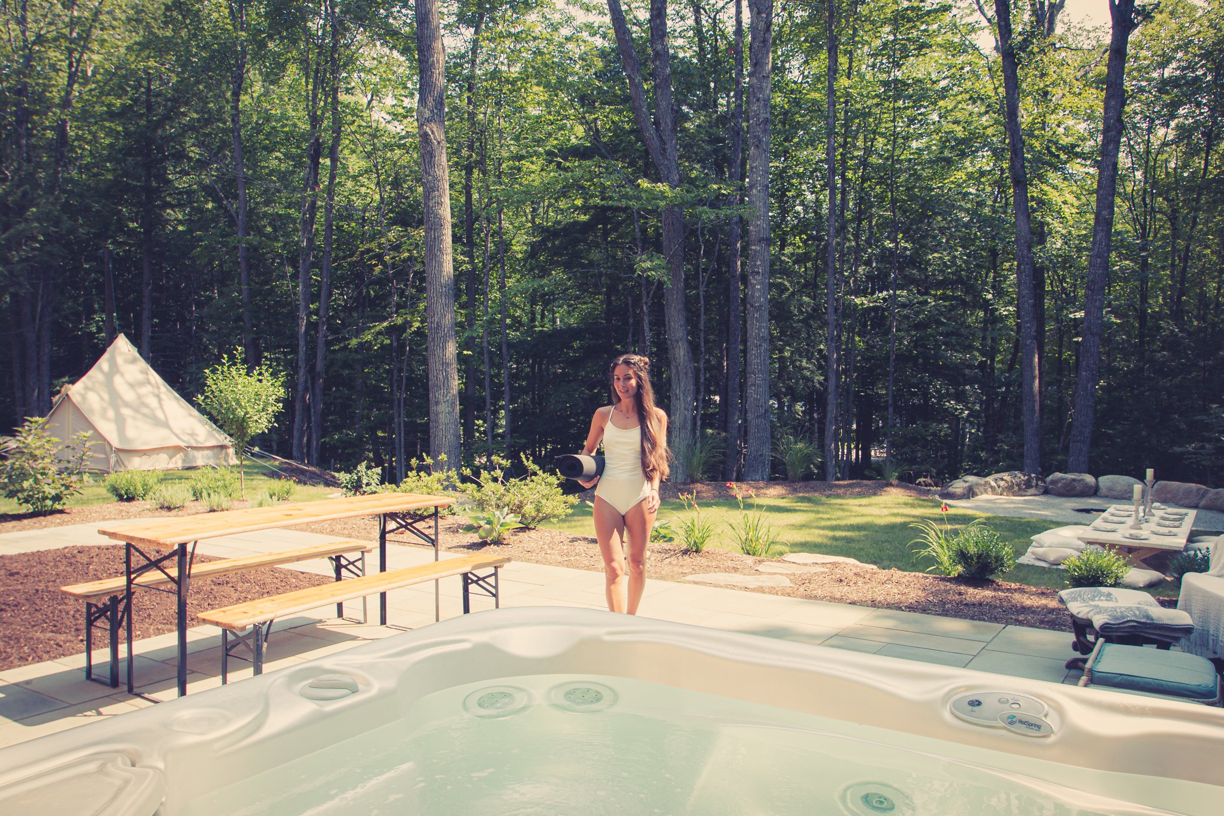 Woman in white swimsuit holding mat, standing near hot tub in forest setting, with picnic benches and tent in the background.