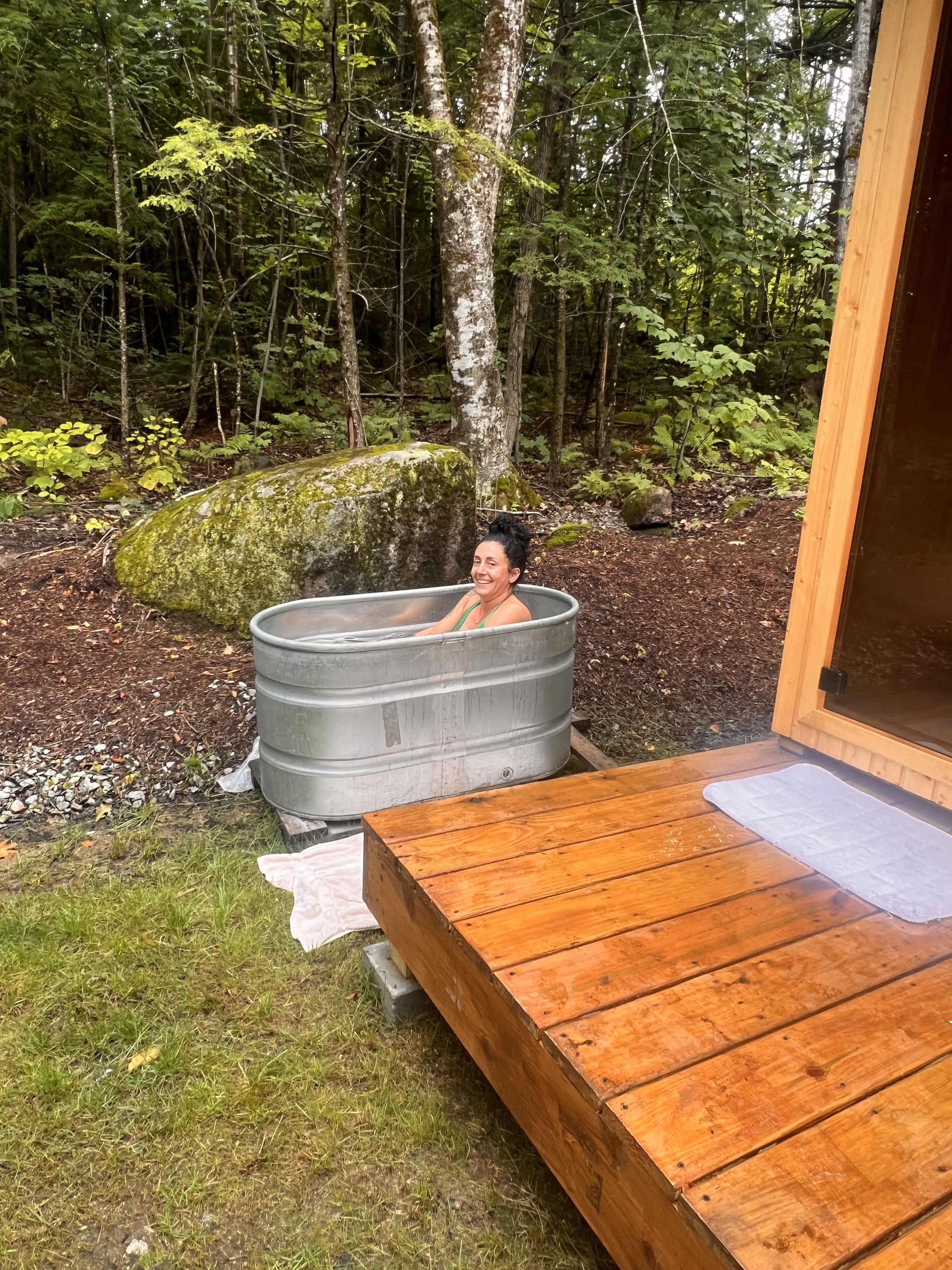 Person relaxing in metal tub in forest near wooden deck