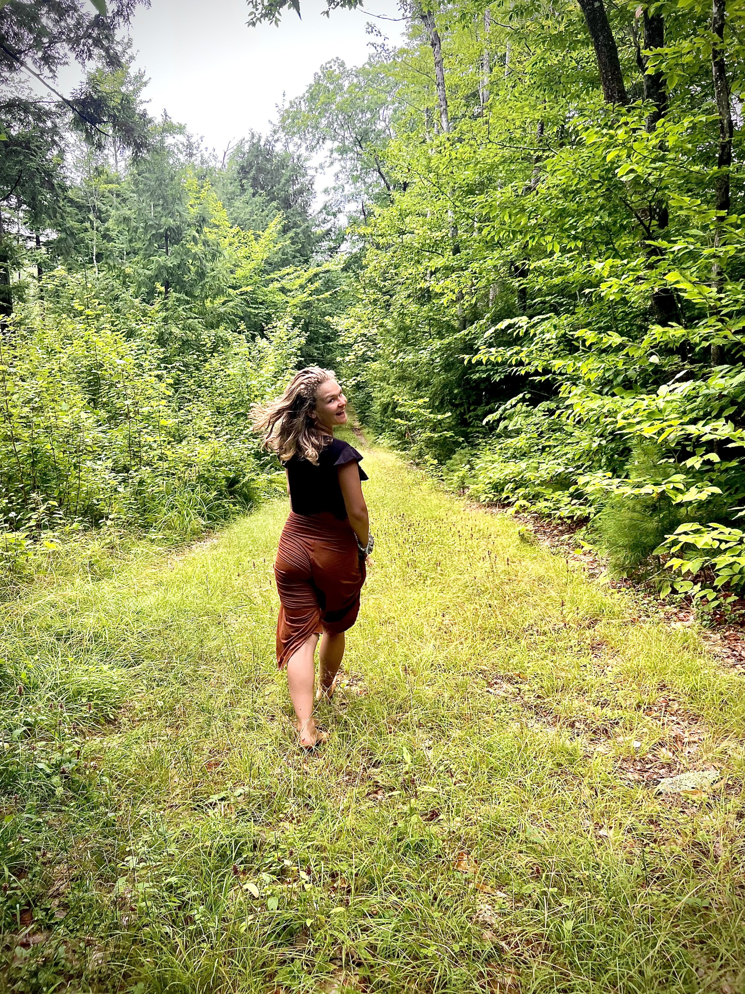 Person walking on grassy path in forest, surrounded by lush green trees, wearing a black top and brown skirt.