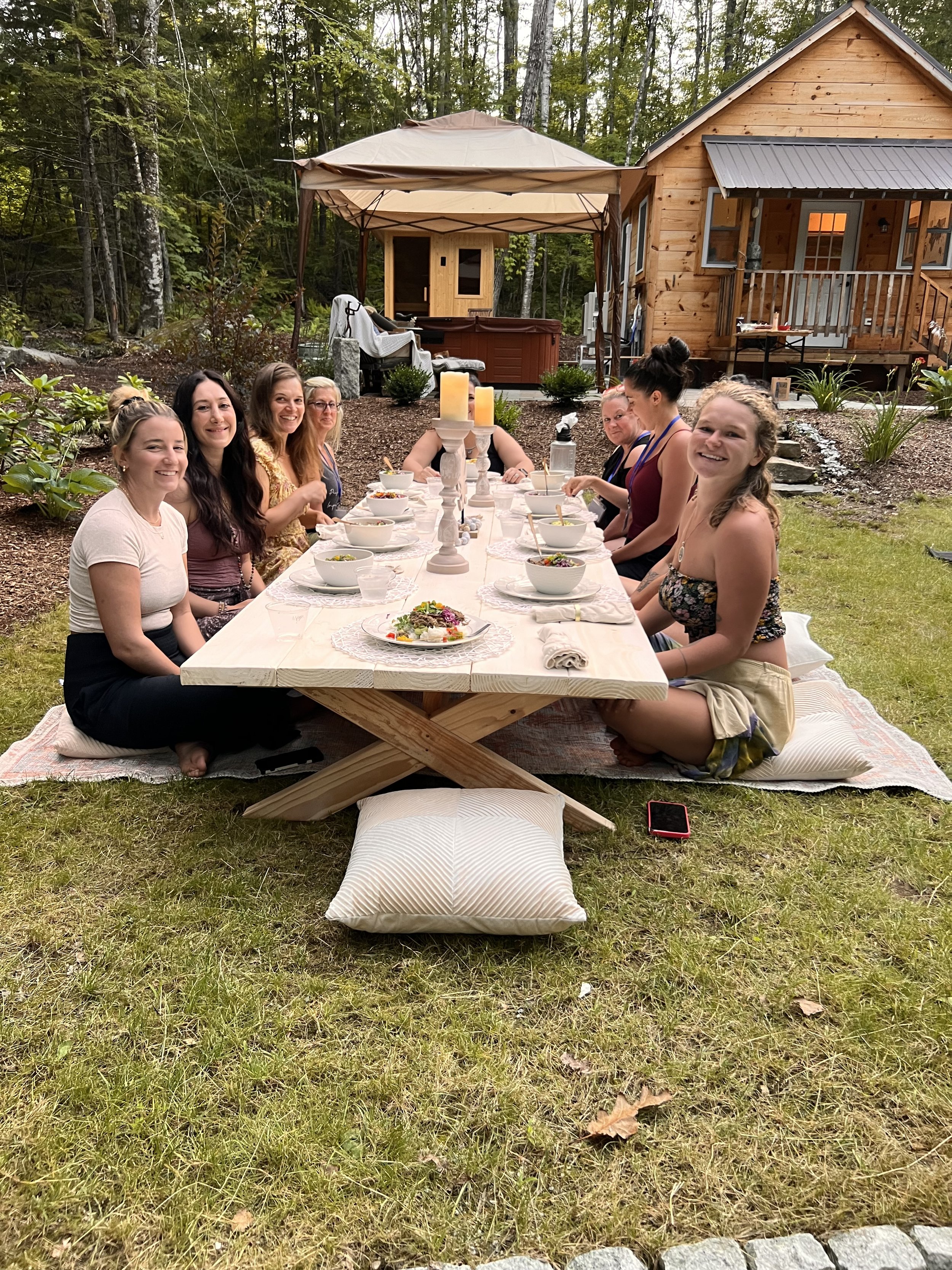 Group of women sitting at an outdoor picnic table set with food, with a wooden cabin and canopy in the background.