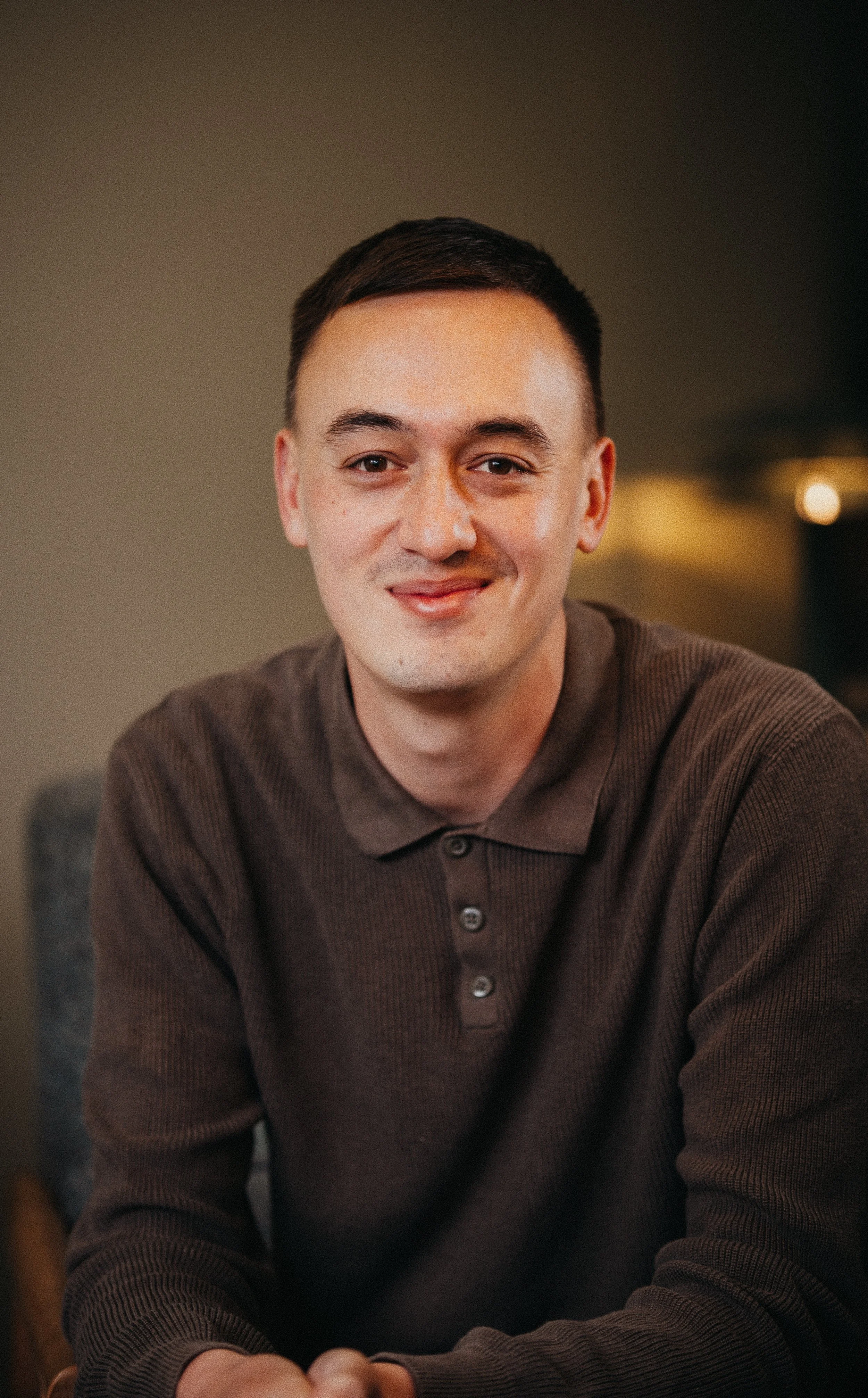 A young man with short dark hair, fair skin, and wearing a brown button-up shirt, smiling softly while sitting indoors with a blurred background.