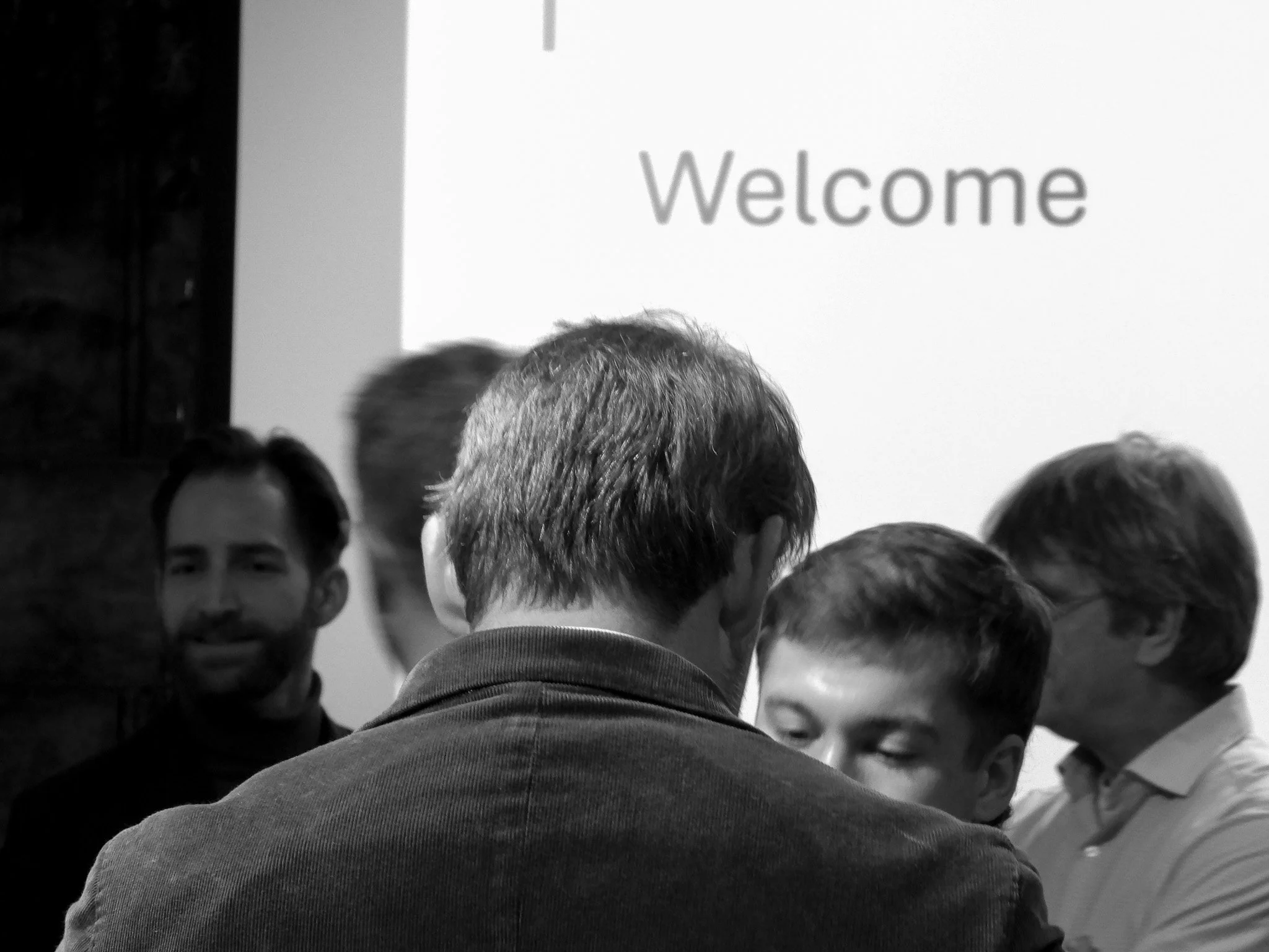 A group of people gathered closely together, facing a white screen or wall with the word 'Welcome' displayed, in an indoor setting.