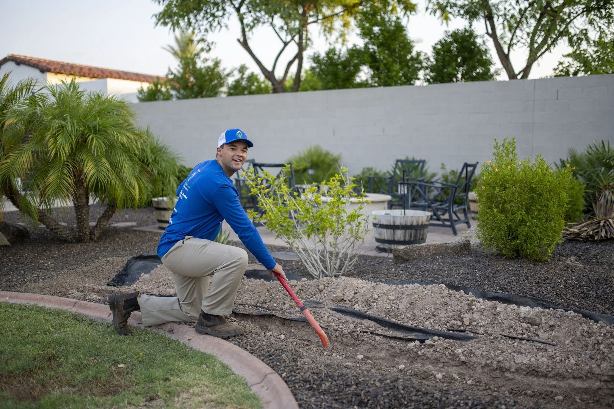 A man kneeling on the ground with one knee, using a small gardening shovel in a landscaped backyard, with lush plants and a patio with chairs and tables in the background.