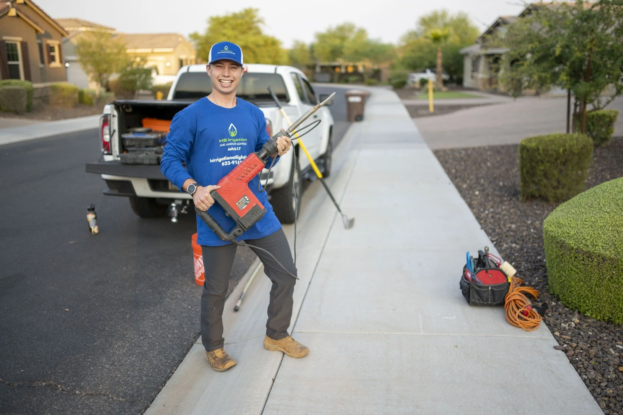 A man in a blue shirt and cap holding a large red power tool, smiling on a sidewalk in front of a pickup truck, with tools and equipment nearby, working outside in a residential neighborhood.