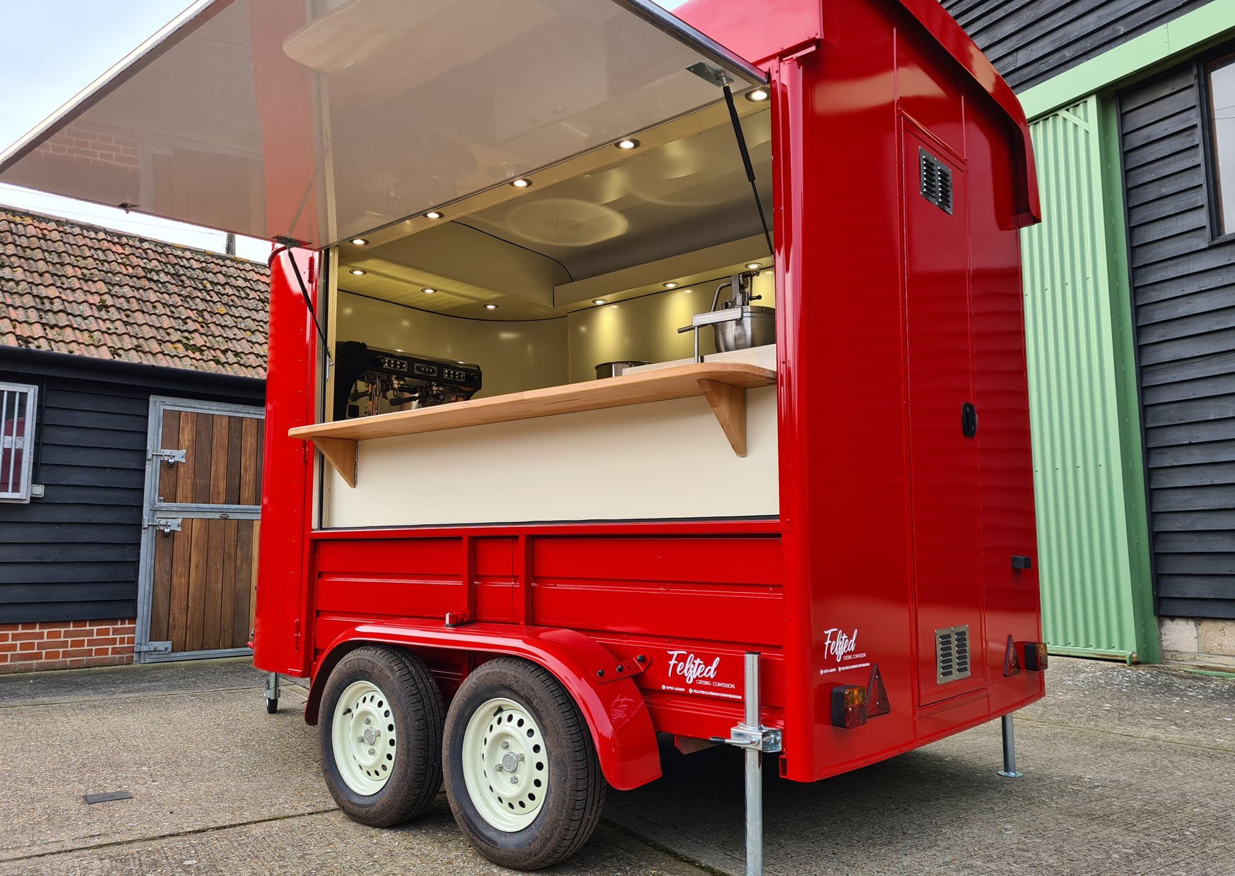 Red mobile coffee trailer with serving counter and espresso machine, parked on concrete surface near barn.