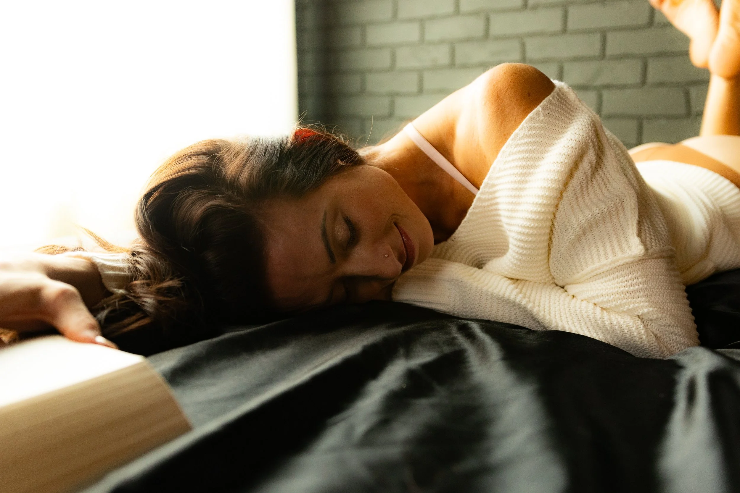 A woman lies on a bed with her head resting softly on black sheets, wearing a cozy off-the-shoulder cream sweater and a light bra strap visible. She smiles gently with her eyes closed as window light glows behind her against a gray brick wall.