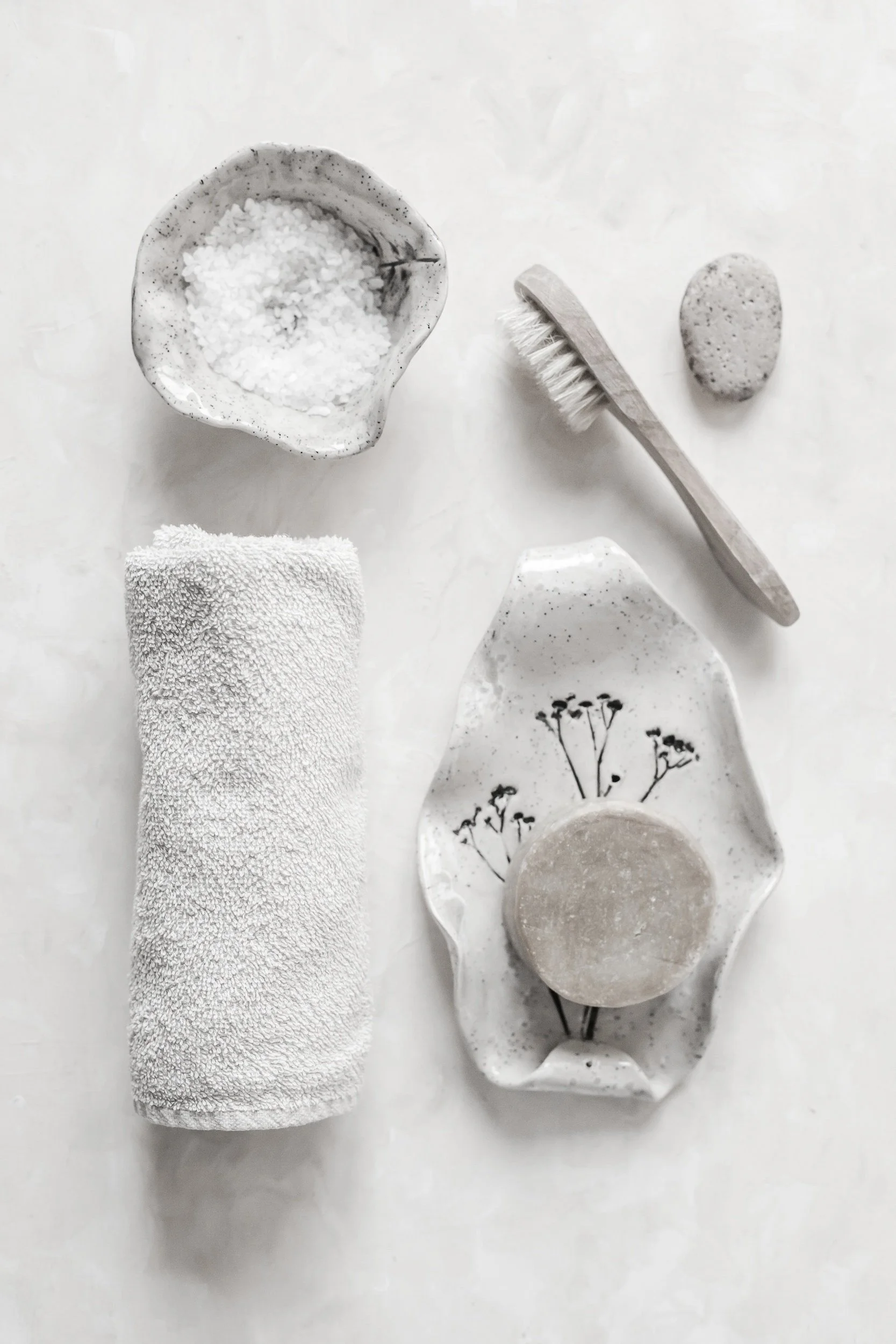 A collection of minimalist bath essentials including a rolled white towel, a ceramic dish with a soap bar, a scrub brush, a pumice stone, a small bowl with bath salts, and a stone, all arranged on a white surface.