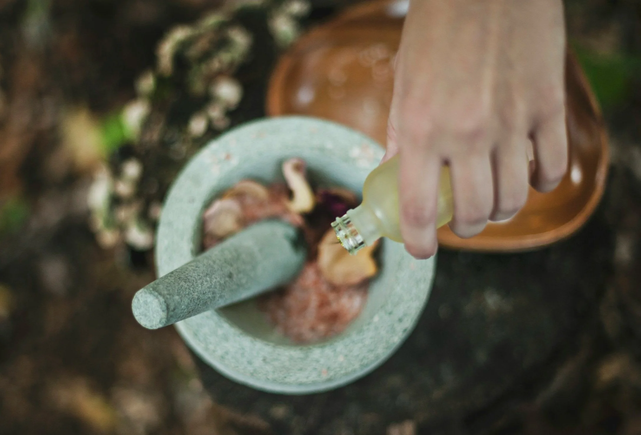 A person pouring liquid into a stone mortar with a pestle, with a wooden tray and blurred natural background nearby.