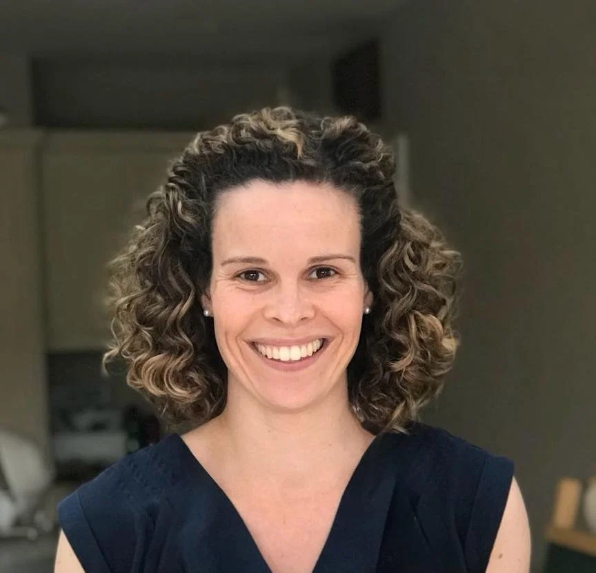 Image of Amanda, she is smiling to the camera and proudly displaying a clay vase she has made, she is wearing an apron and there are other pottery pieces and clay in the background
