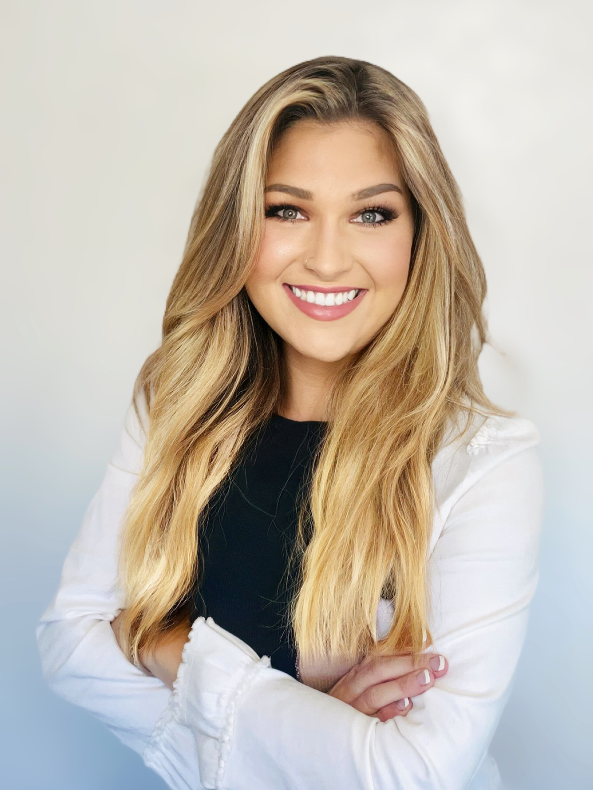 A young woman with long, wavy blonde hair, smiling with her arms crossed, wearing a black top and white jacket against a light background.