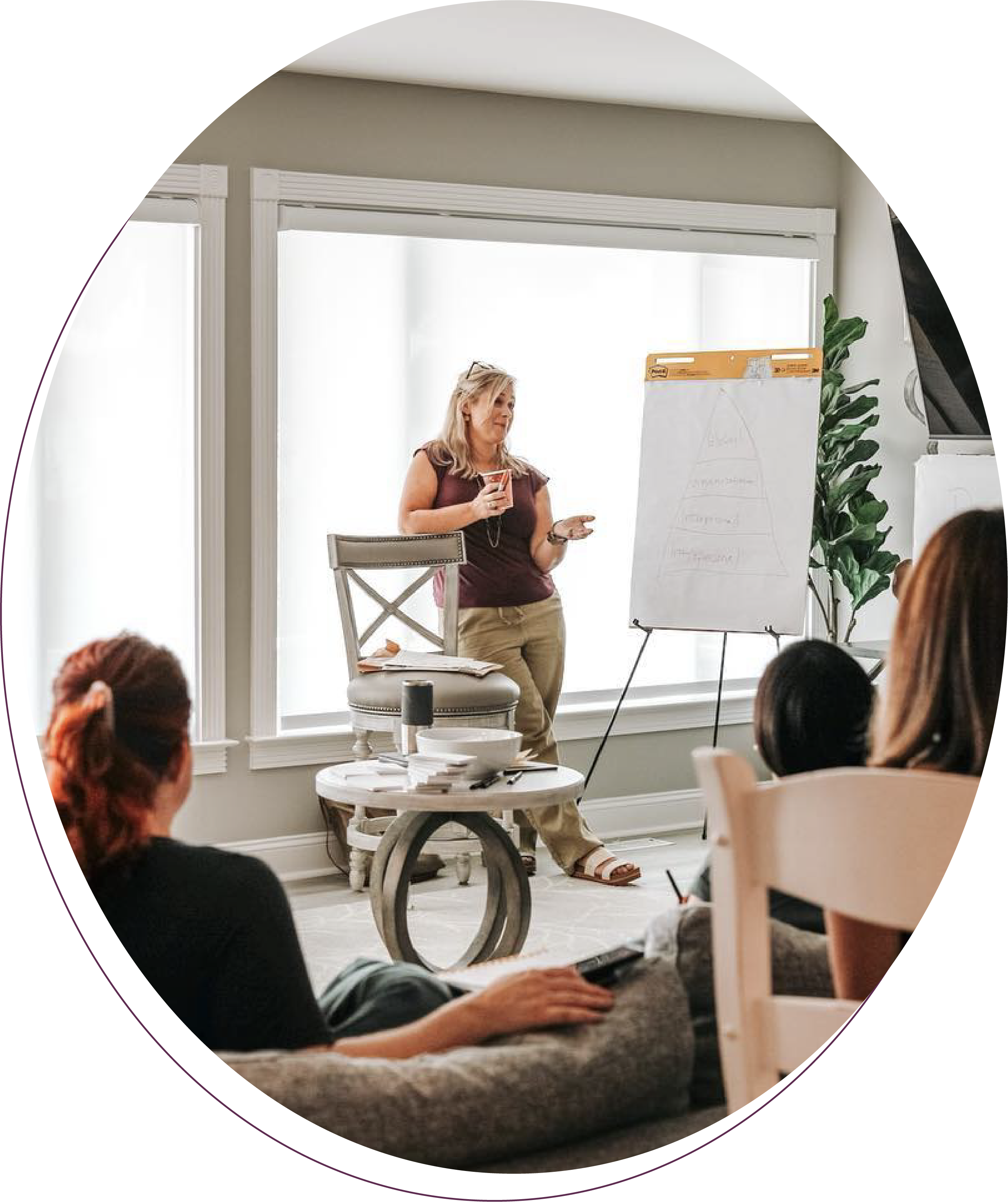 A woman giving a presentation to a small group in a bright room with large windows, a whiteboard with writing, and a round table with papers, bowls, and a water bottle.