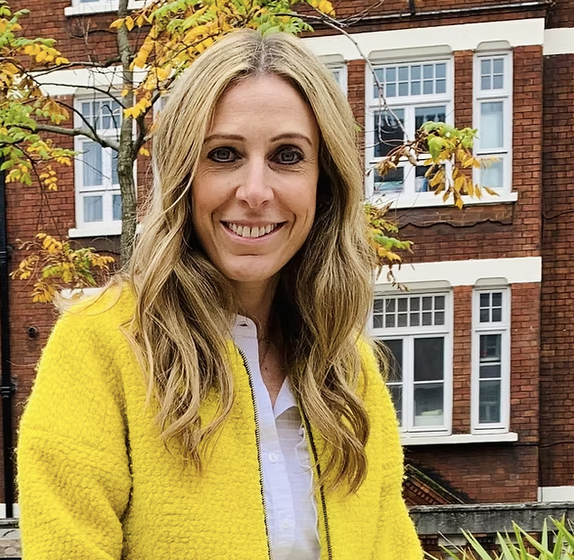 A woman with long blonde hair smiling outdoors, wearing a yellow jacket and white shirt, with a red brick building and trees with fall leaves in the background.