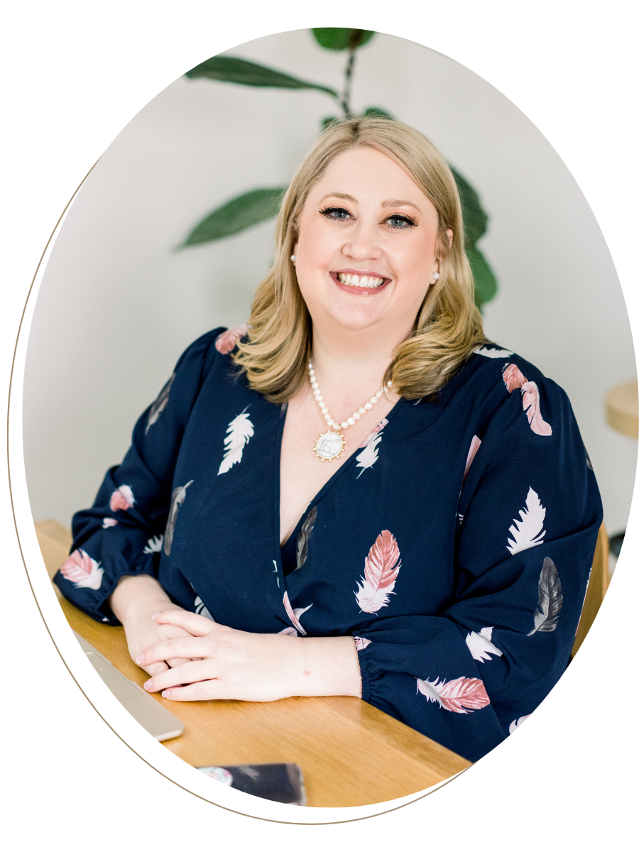 A woman with blonde hair, wearing a navy blue blouse with a feather pattern, pearl necklace, pearl earrings, smiling at the camera, sitting at a wooden table with a laptop and phone. Background has a green leafy plant.