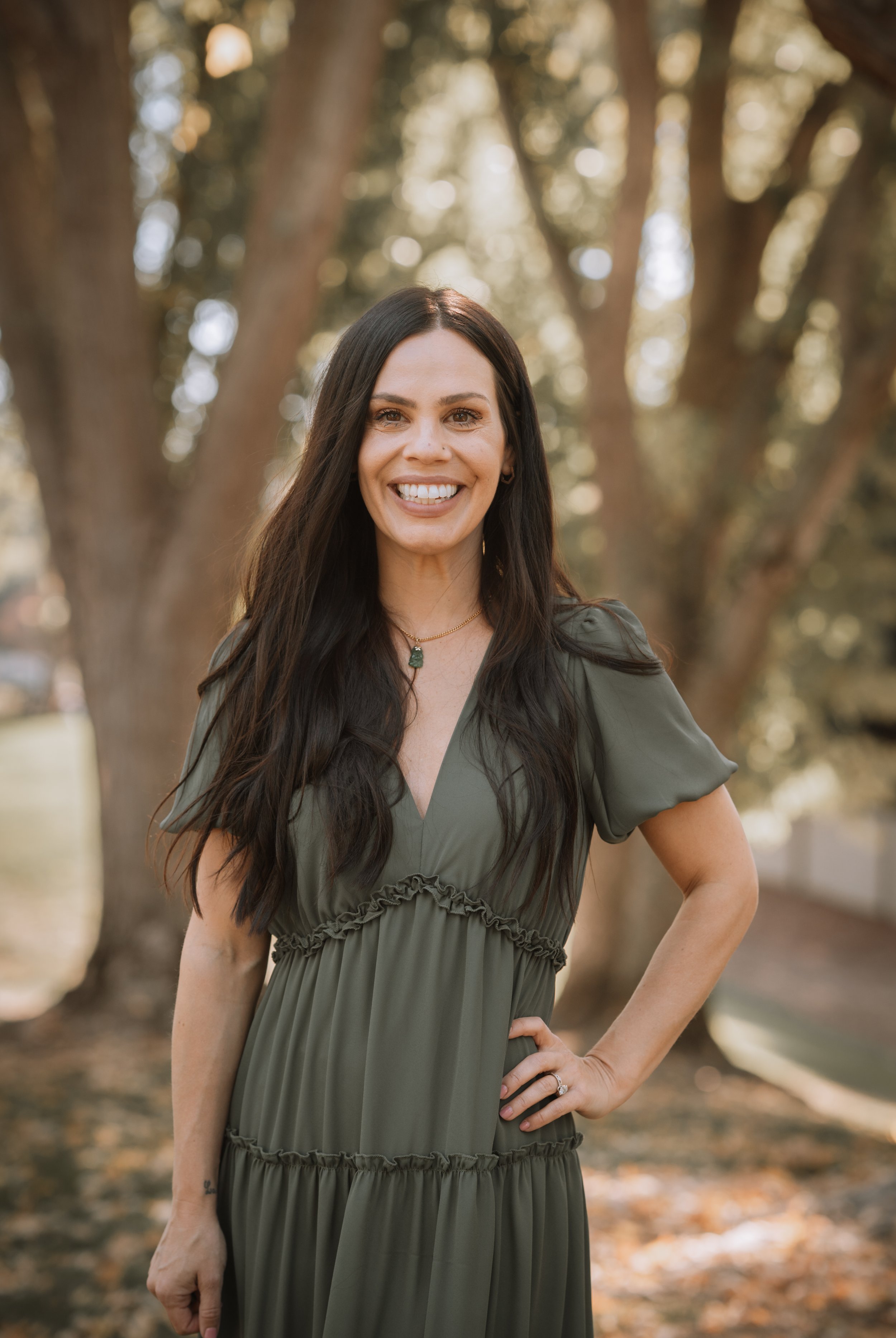 A woman with long dark hair smiling outdoors in front of trees, wearing a green dress.