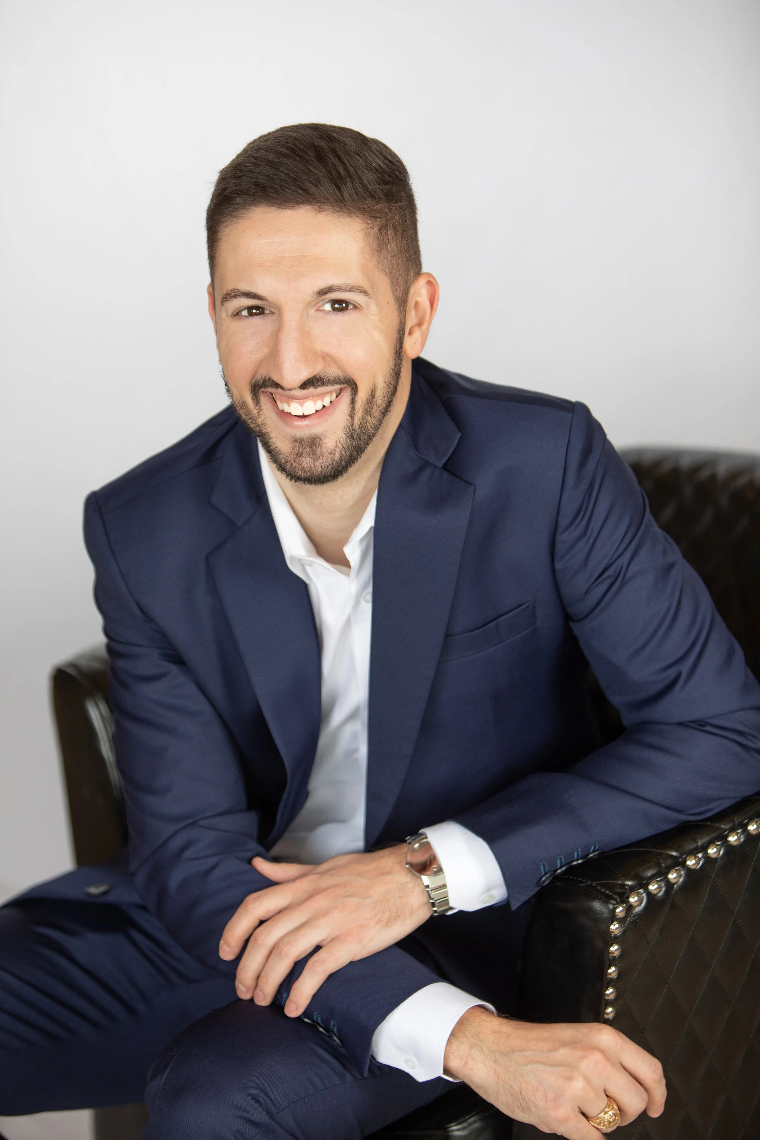 A man in a navy blue suit with a white shirt sitting on a black leather chair, smiling.