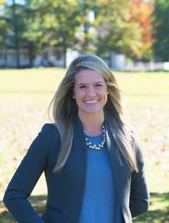 A smiling woman with shoulder-length blonde hair, wearing a navy blazer, a blue top, and a pearl necklace, outdoors on a sunny day with green grass and trees in the background.