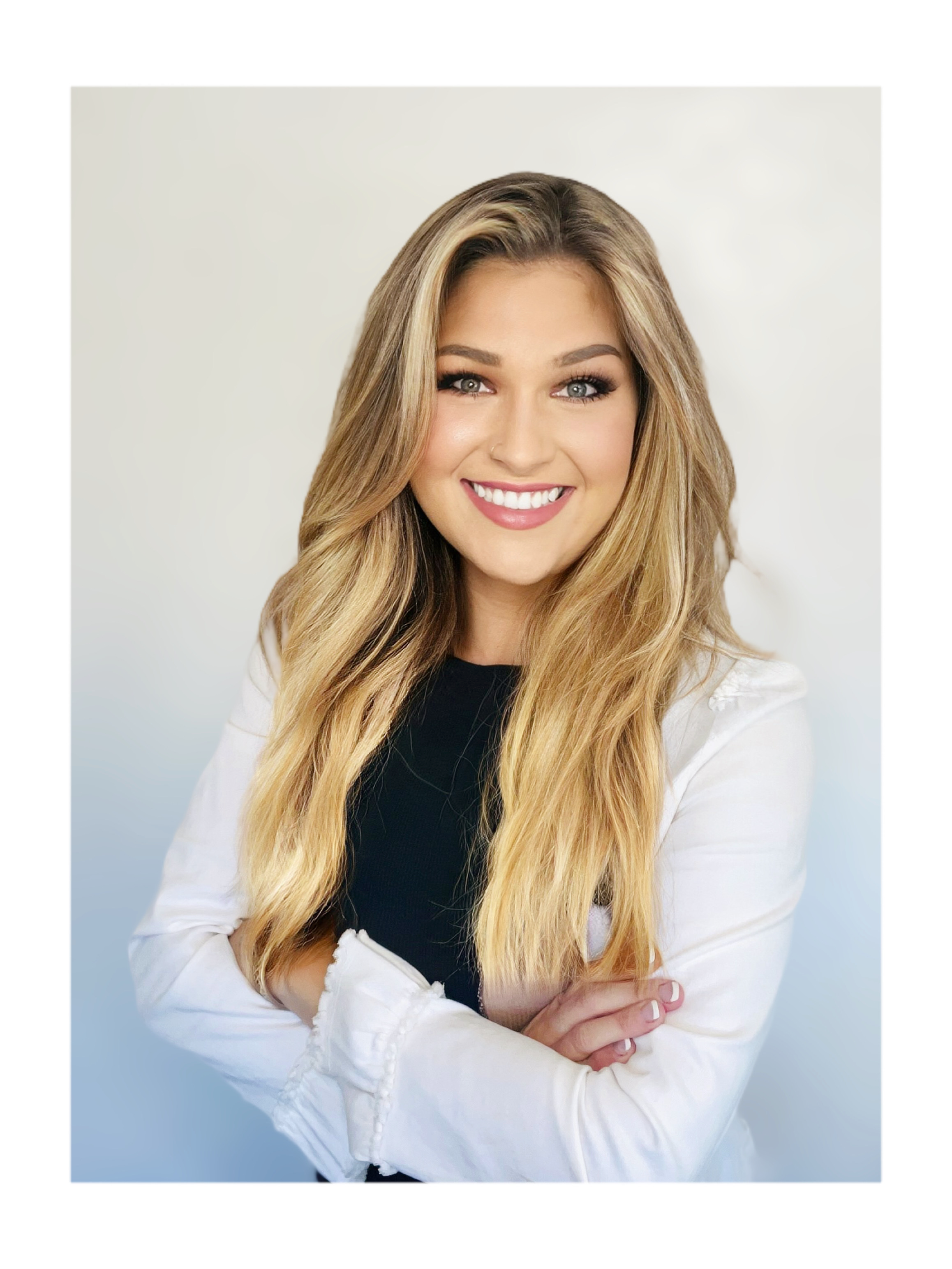 Portrait of a young woman with long blonde hair, smiling, wearing a white and black top, with arms crossed, indoor studio setting.