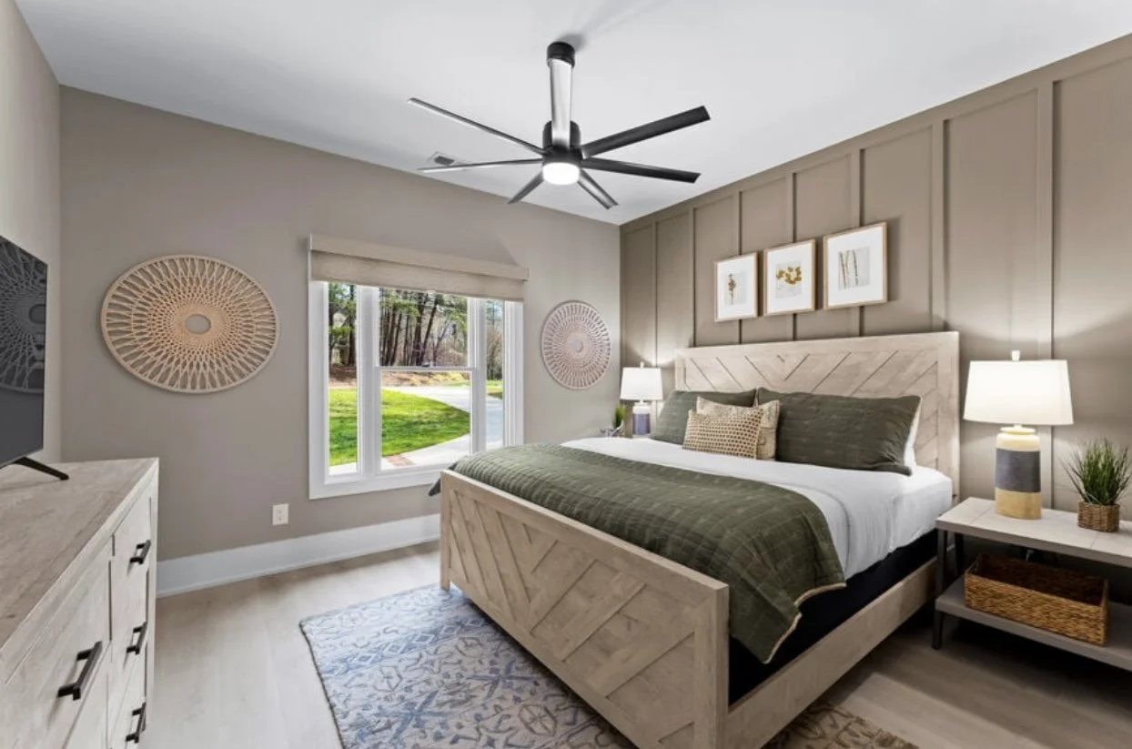 Bedroom with beige walls, wooden furniture, green bedding, and artwork above the bed. Sunlight coming through a window showing a grassy yard outside.