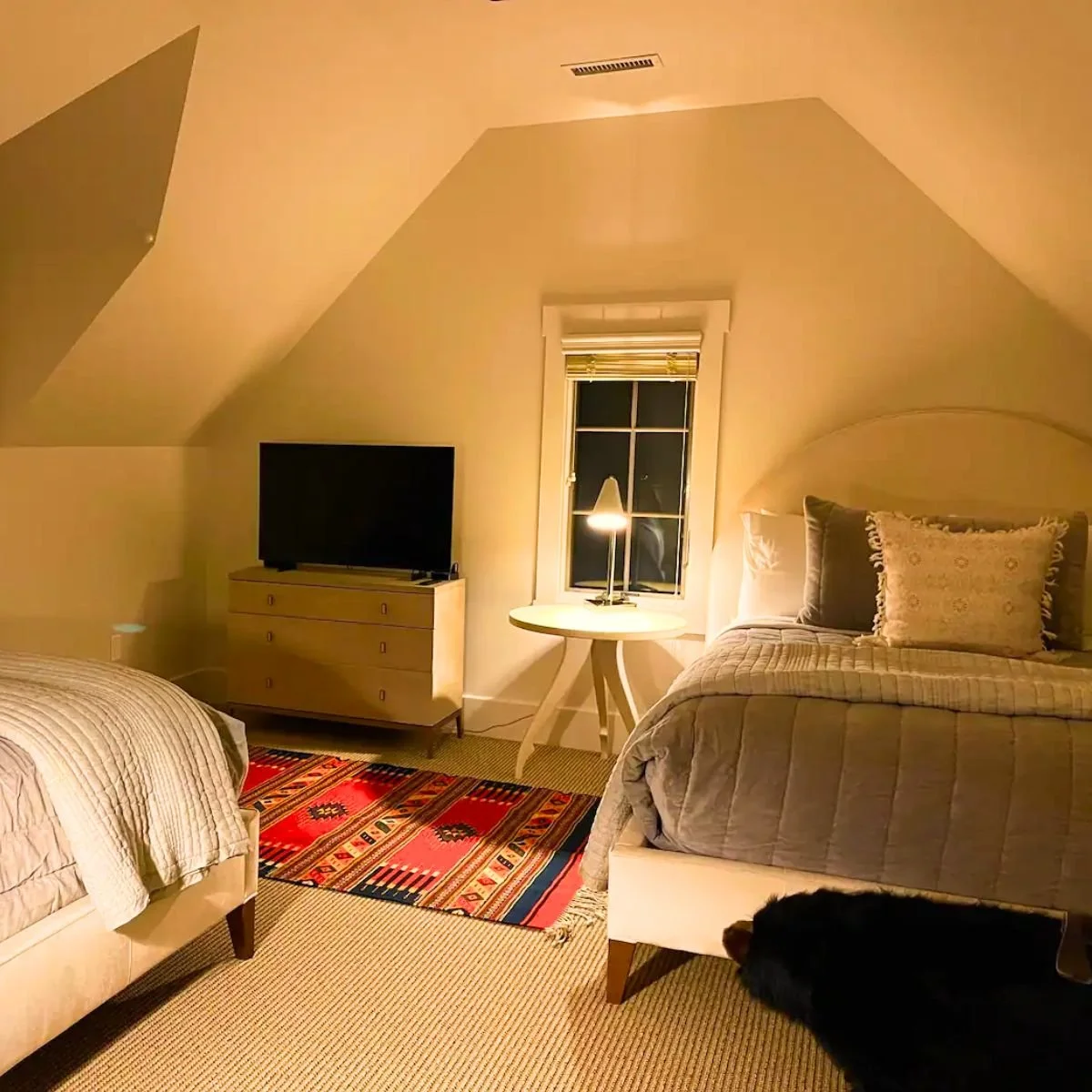 A cozy bedroom with sloped ceiling, two beds, a small round table with a lamp, a television on a dresser, a window with blinds, and a colorful rug on the carpeted floor. A black dog is partially visible under one of the beds.