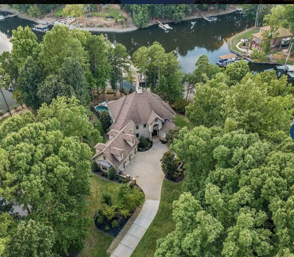 A large house with a curved driveway surrounded by lush green trees, located near a river with boats and dock space.