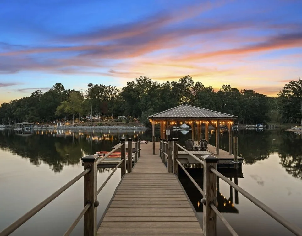 Dock extending over calm lake water during sunset with trees and sky in the background, and a covered seating area with string lights at the end of the dock.