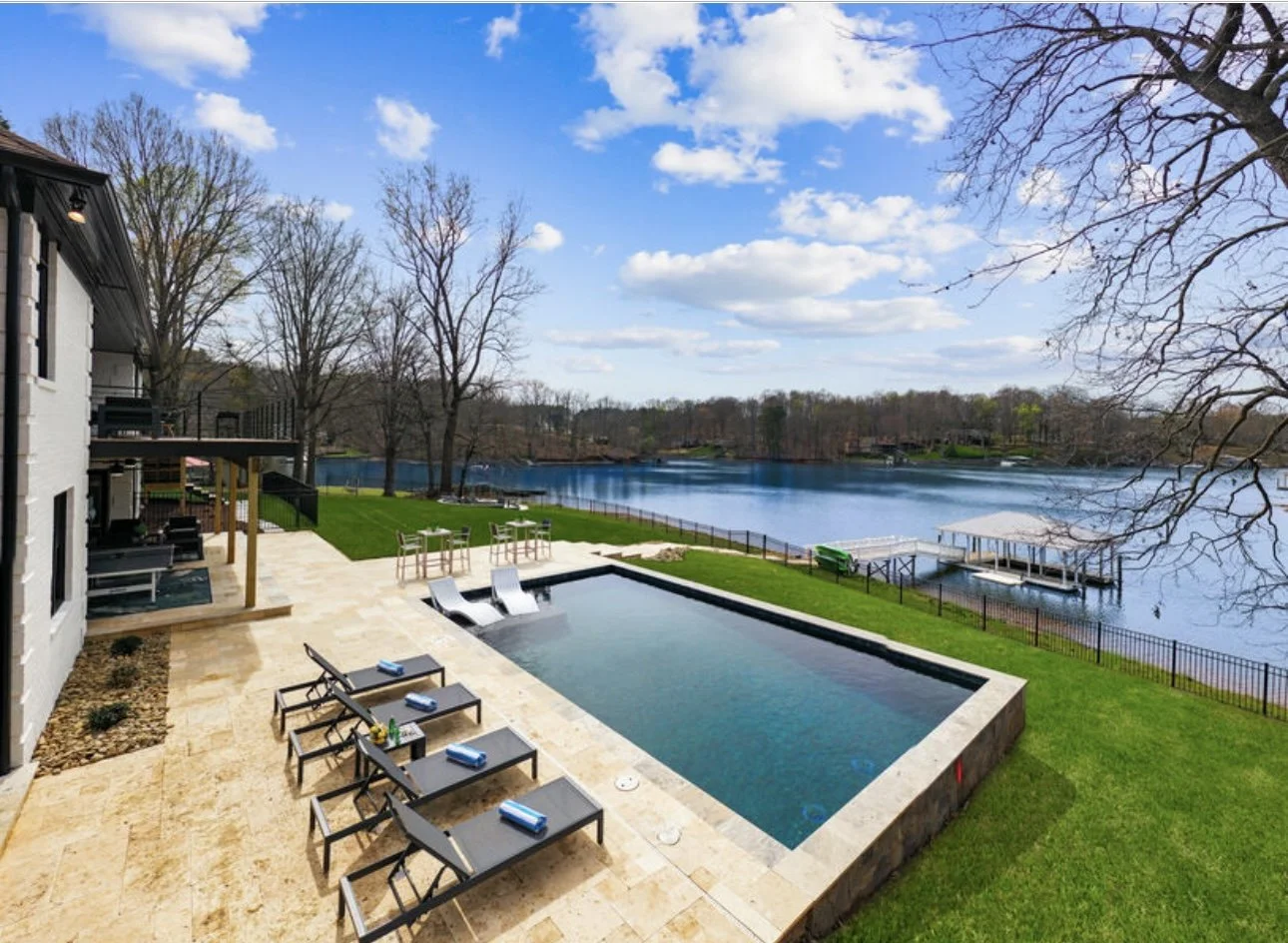 A backyard with a poolside lounge area, a small pool with waterfalls, a grassy yard, a lake with a dock, and trees with bare branches under a partly cloudy sky.