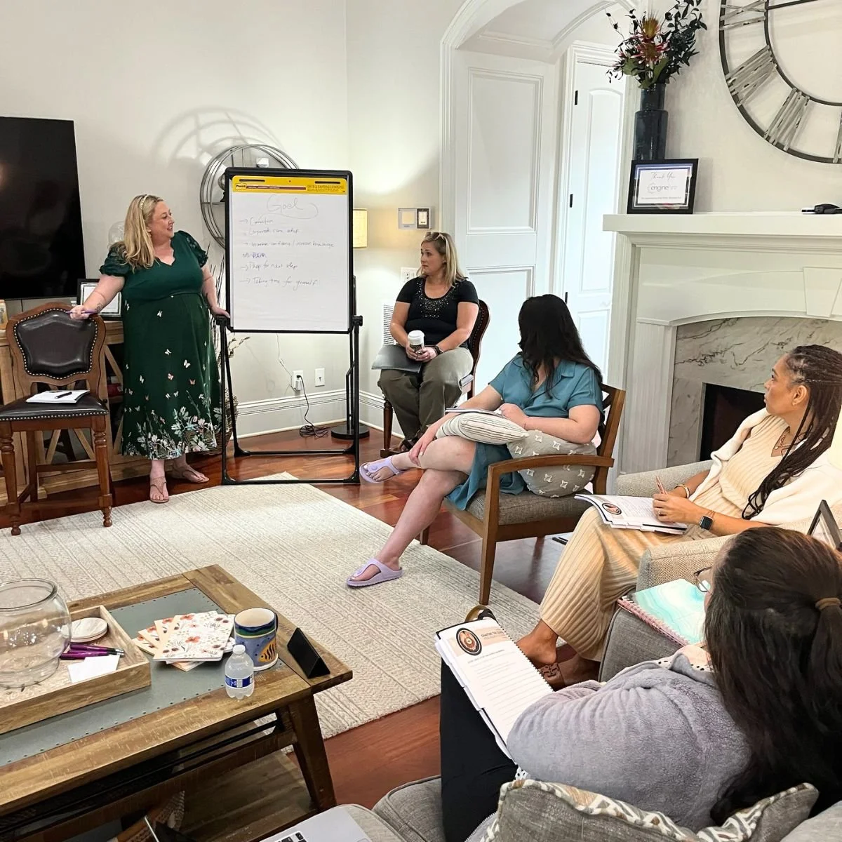 Women participating in a group meeting or workshop in a cozy living room with white walls, a fireplace, and a clock on the wall, with one woman standing and presenting notes on a whiteboard while others are seated and listening.