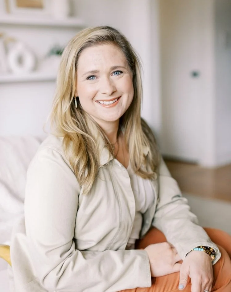 A smiling woman with long blonde hair and blue eyes sitting on a beige couch in a cozy, well-lit room, wearing a light-colored jacket and a bracelet.