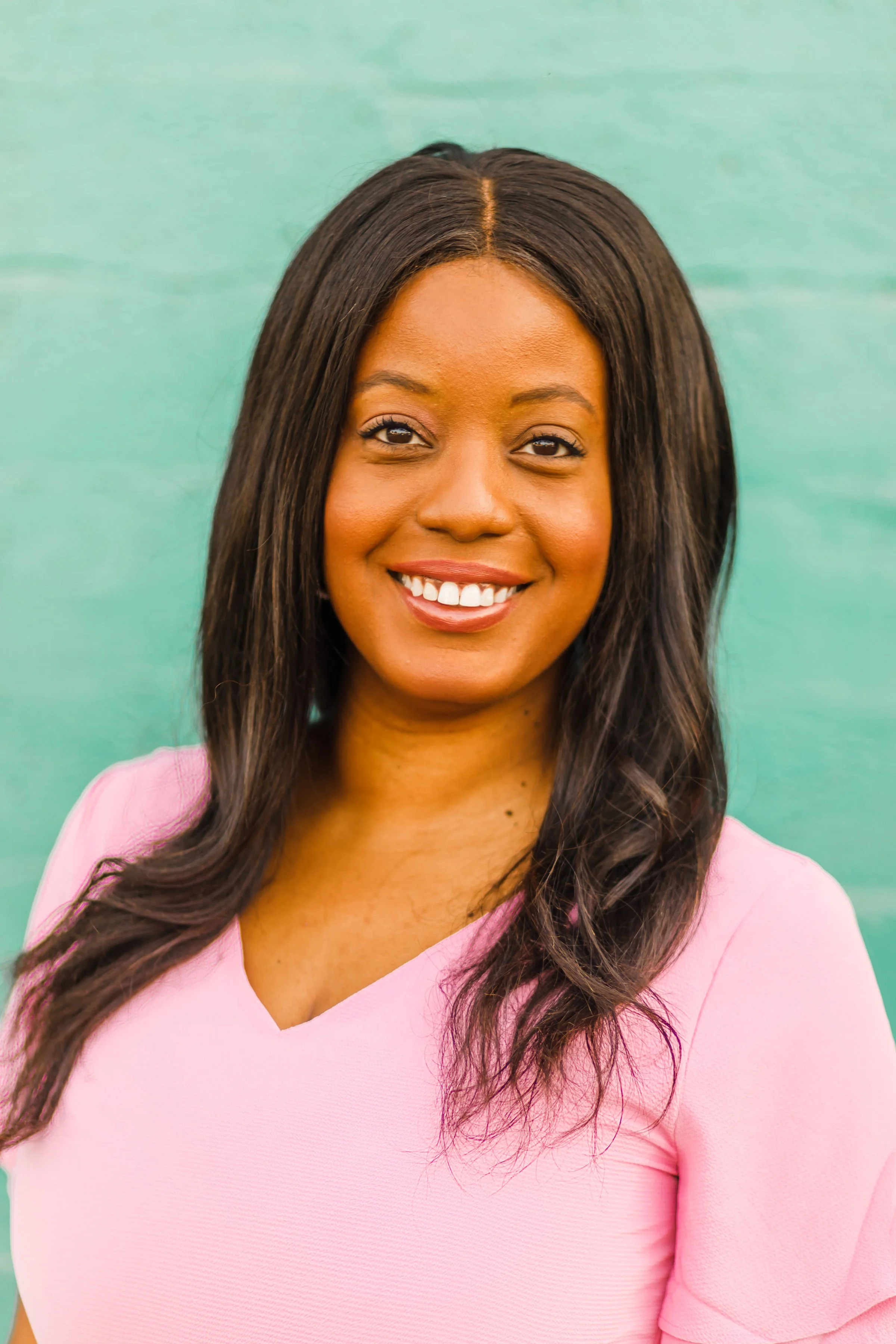 A woman with long dark hair smiling in front of a green background, wearing a pink top.