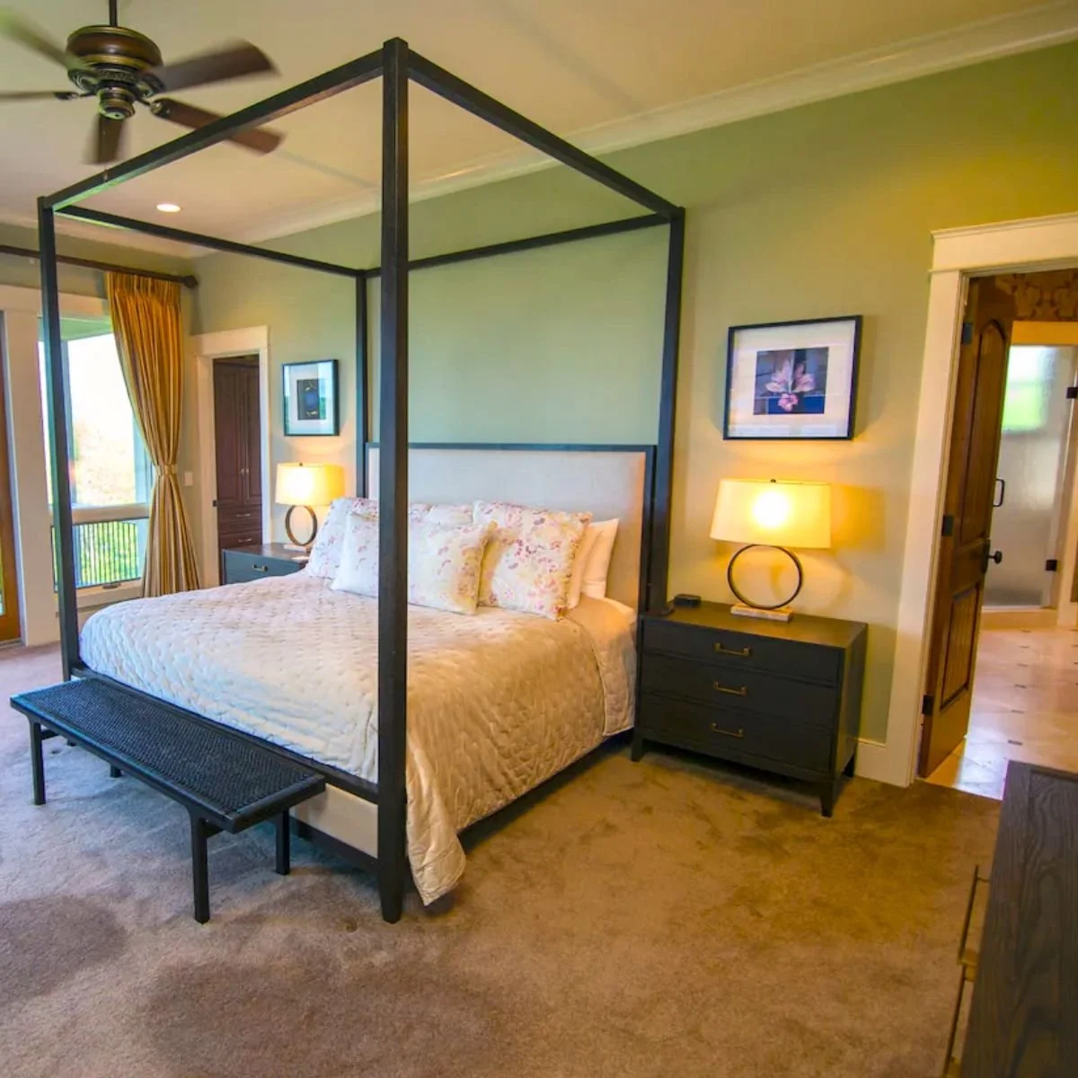 A bedroom with a four-poster bed, bedside tables with lamps, art on the walls, a ceiling fan, and a window with yellow curtains.
