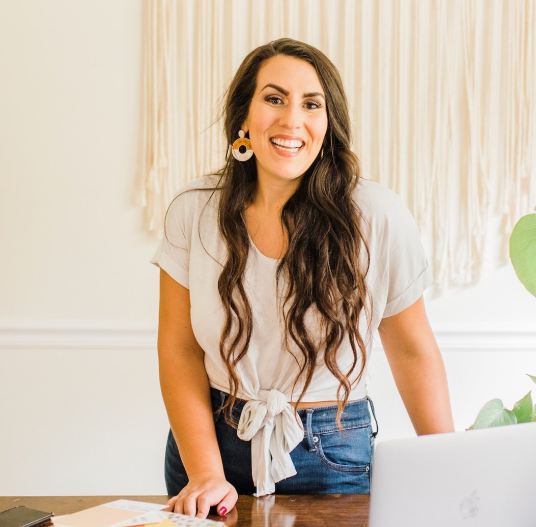 A smiling woman with long wavy brown hair, wearing a white tied-front T-shirt and blue jeans, standing behind a wooden desk with a laptop and papers, in a well-lit room with cream-colored walls and a beige curtain.