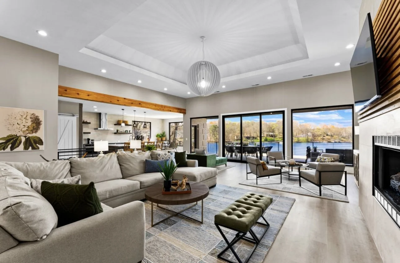 Modern open-concept living room with large windows overlooking a lake, featuring white sectional sofa, armchairs, fireplace, and vaulted ceiling with a modern chandelier.