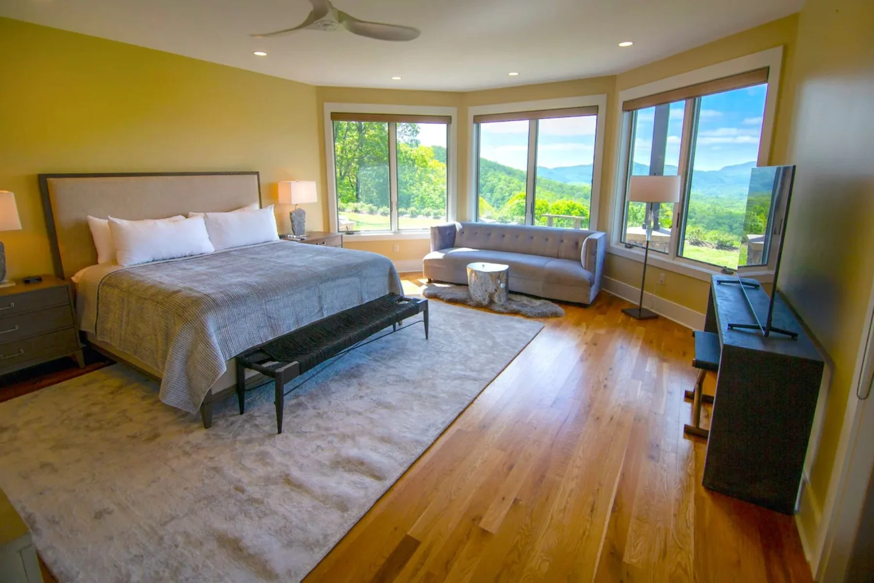Bright bedroom with large windows showing scenic mountain and greenery view, king size bed with neutral bedding, cream sofa near the windows, wooden floor, flat-screen TV, and modern lamps.
