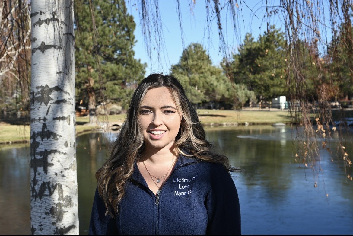 A woman standing outdoors near a tree by a lake on a sunny day.