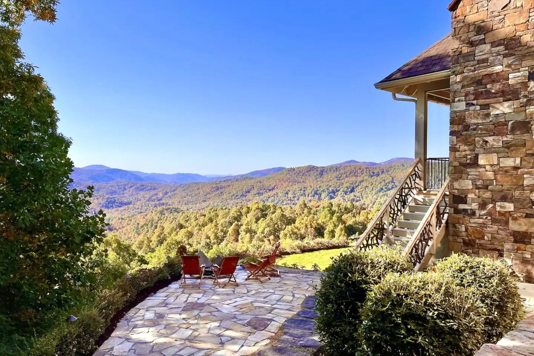 A stone house with stairs leading to a balcony overlooking a scenic mountain landscape with a clear blue sky, and outdoor chairs on a stone patio.