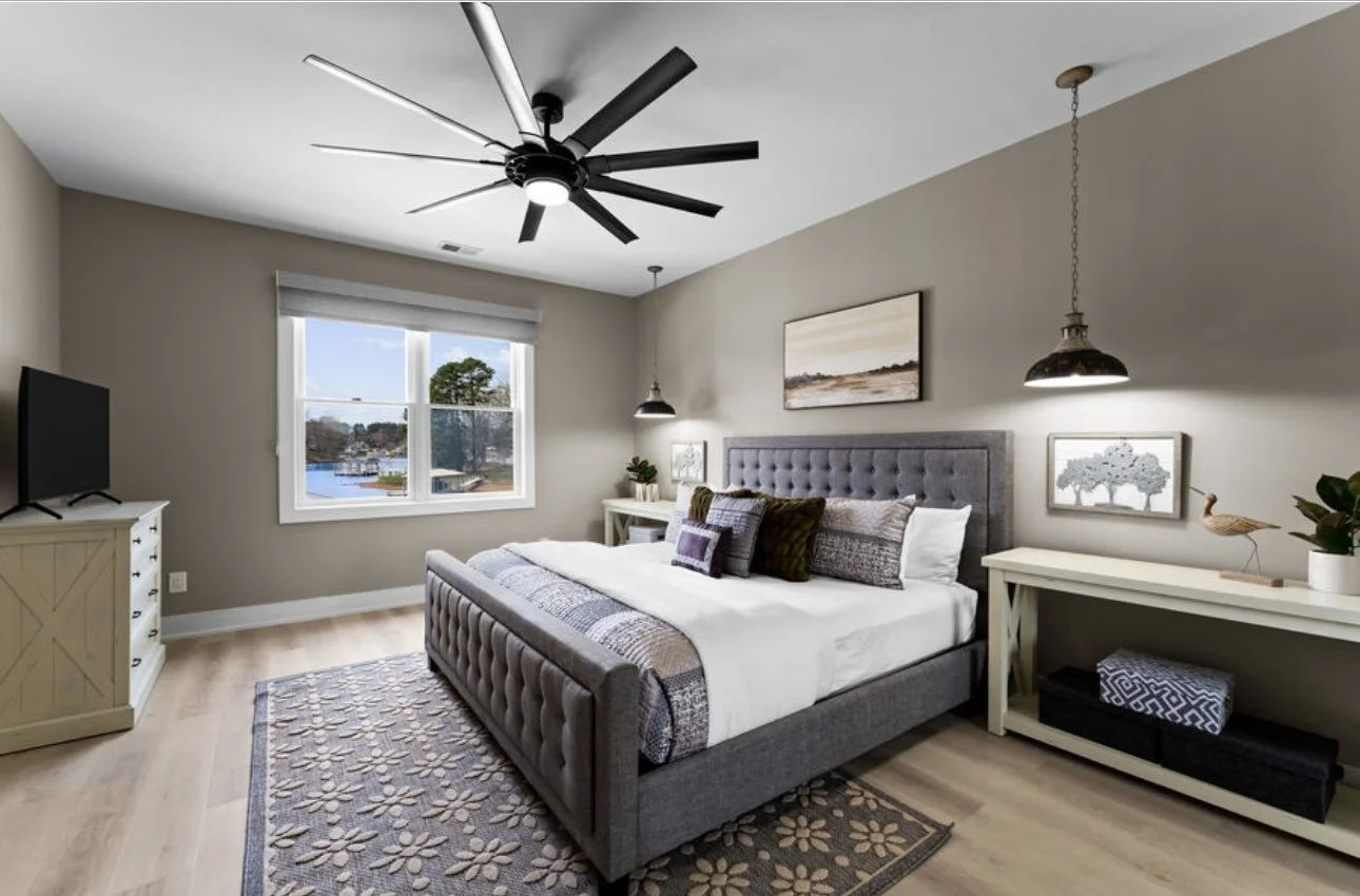 Modern bedroom with gray upholstered bed, beige walls, light wood flooring, window with view of lake and trees, black ceiling fan, two hanging pendant lights, white dresser with TV, nightstands, and decorative artwork and plants.