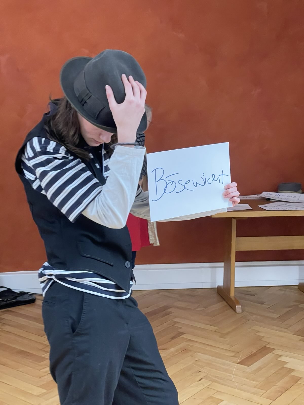 eine junge Frau hält ein Schild in der Hand auf dem "Bösewicht" steht. Mit der anderen Hand fasst sie ihren dunklen Hut an und hat den Kopf leicht gebeugt