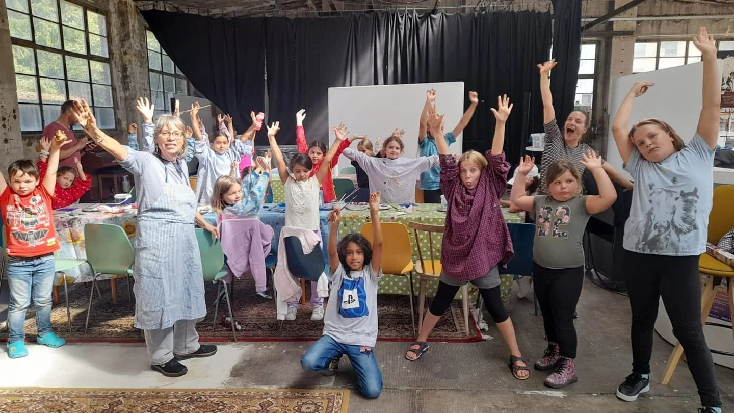 Die Kids, Dani und Simone strecken die Ärme in die Lüfte. Sie stehen vor einem Tisch an dem mit Farbe gewerkelt wurde